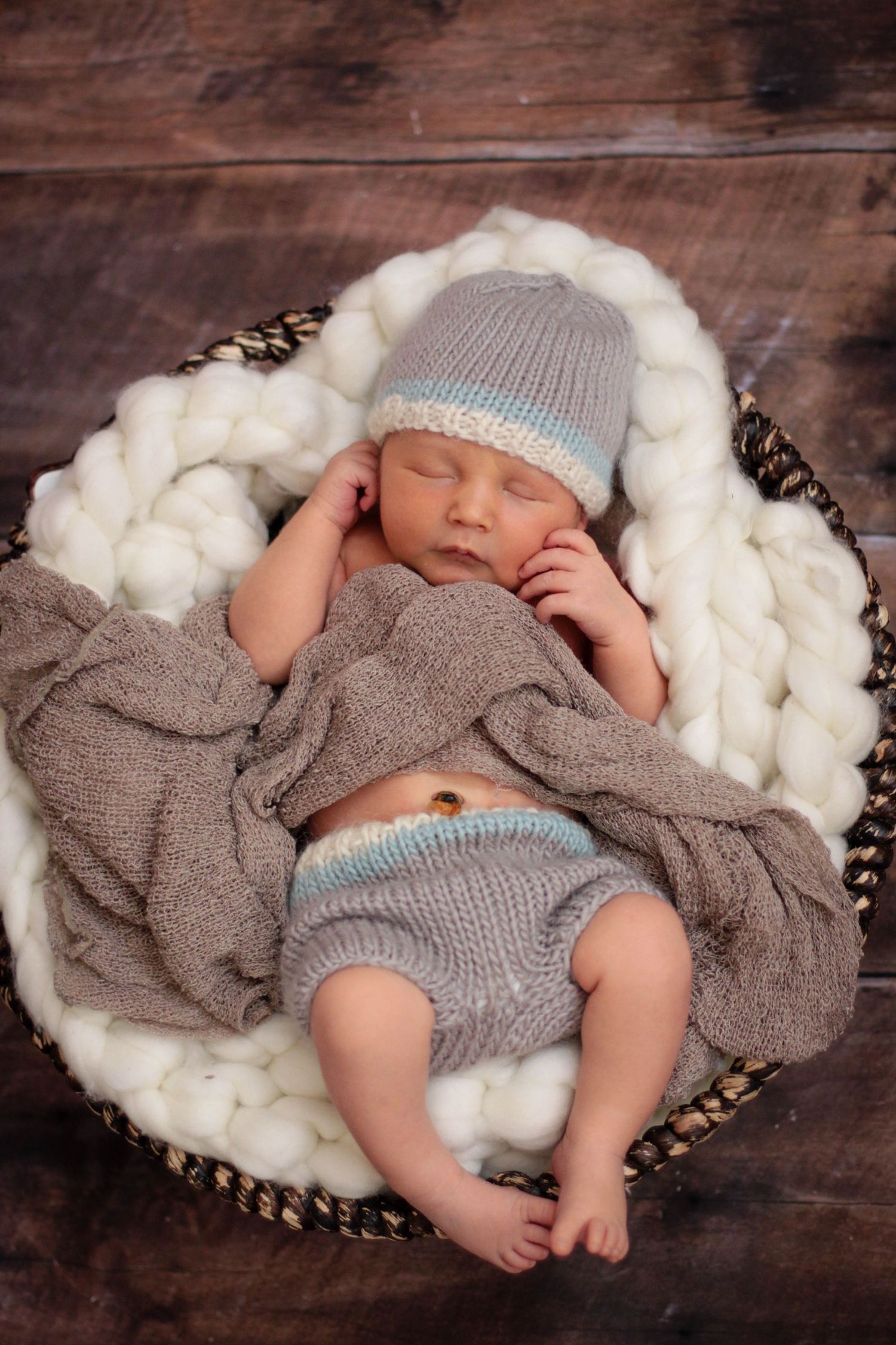 Newborn wearing gray knit outfit sleeping on fluffy white wool backdrop.