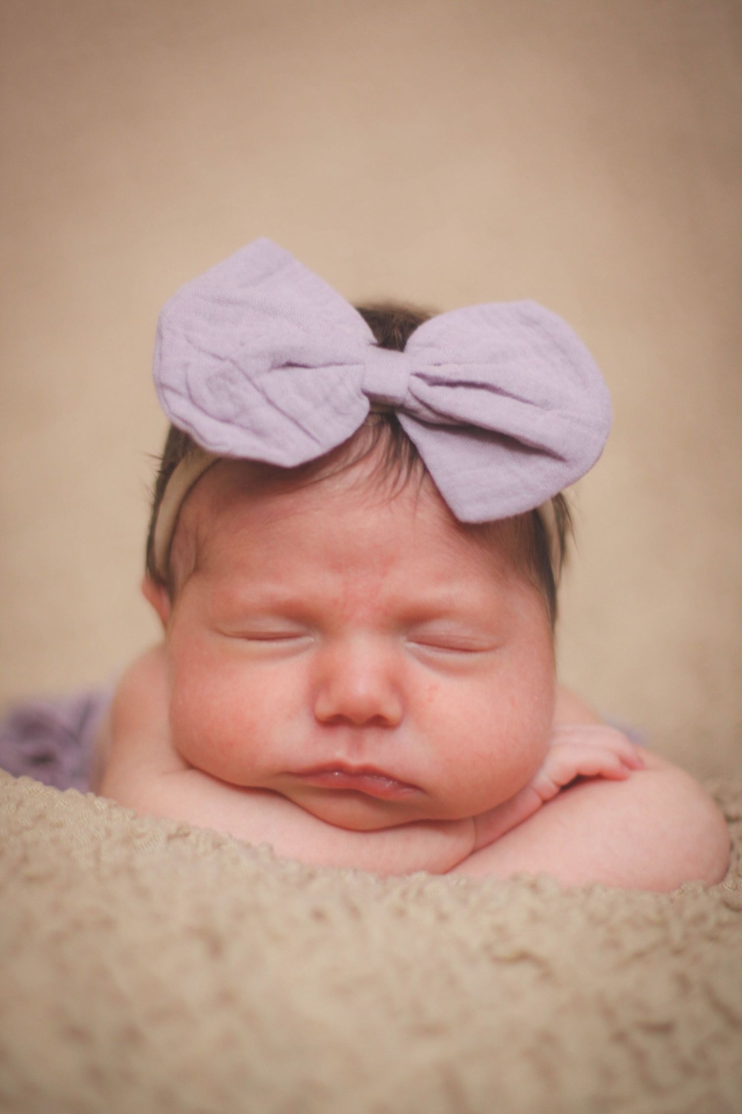 Sleeping baby with lavender bow headband on beige textured background.