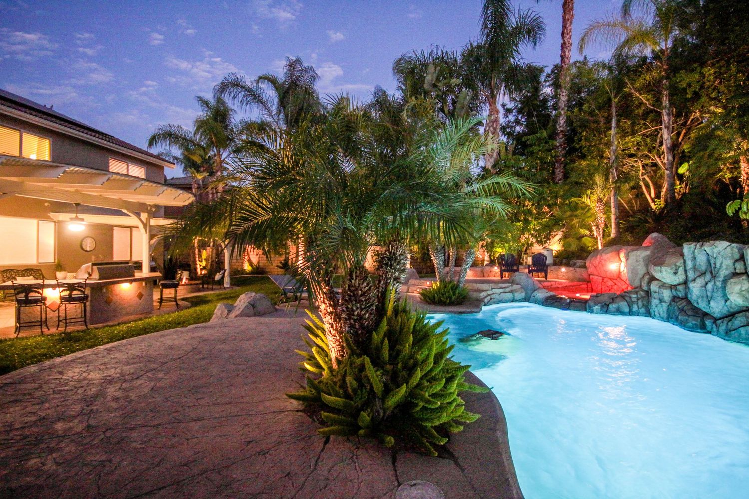 Luxurious backyard pool area illuminated at dusk with tropical landscaping and ambient lighting.