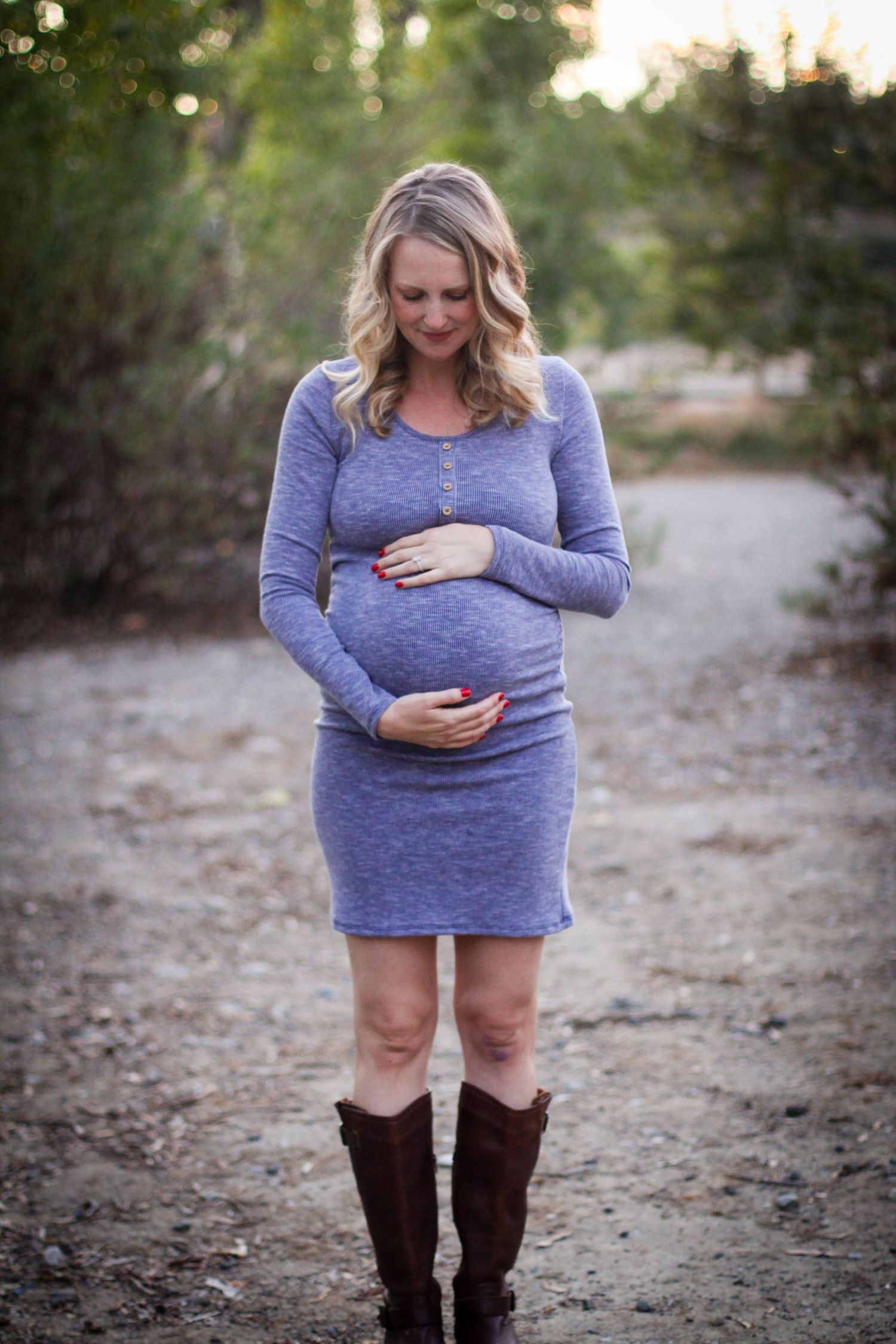 A woman in a purple sweater and brown boots stands on a dirt path during a maternity photoshoot outdoors.