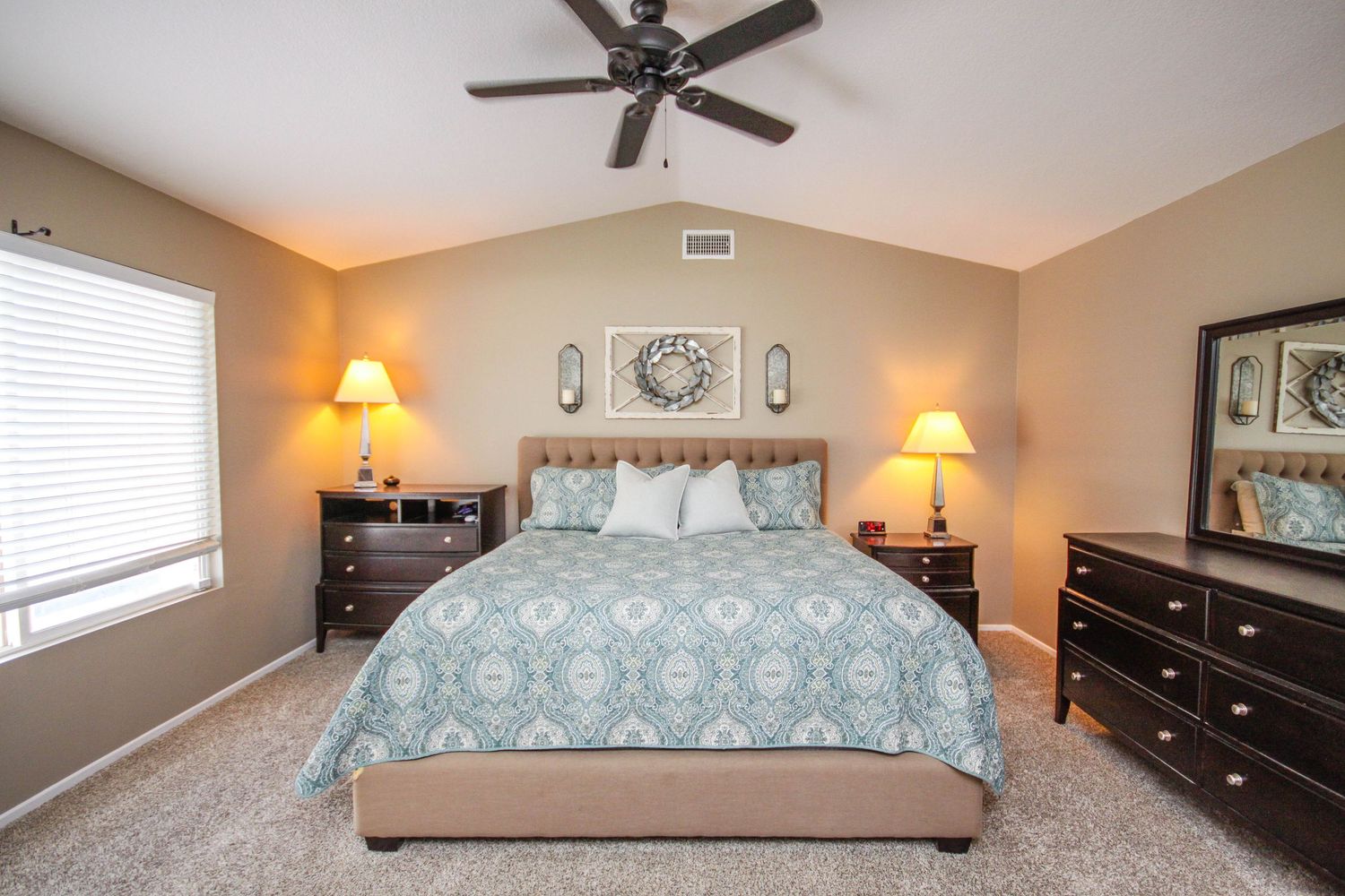 A panoramic view of a cozy bedroom with ceiling fan, wall art, and dark wood furniture in neutral tones.