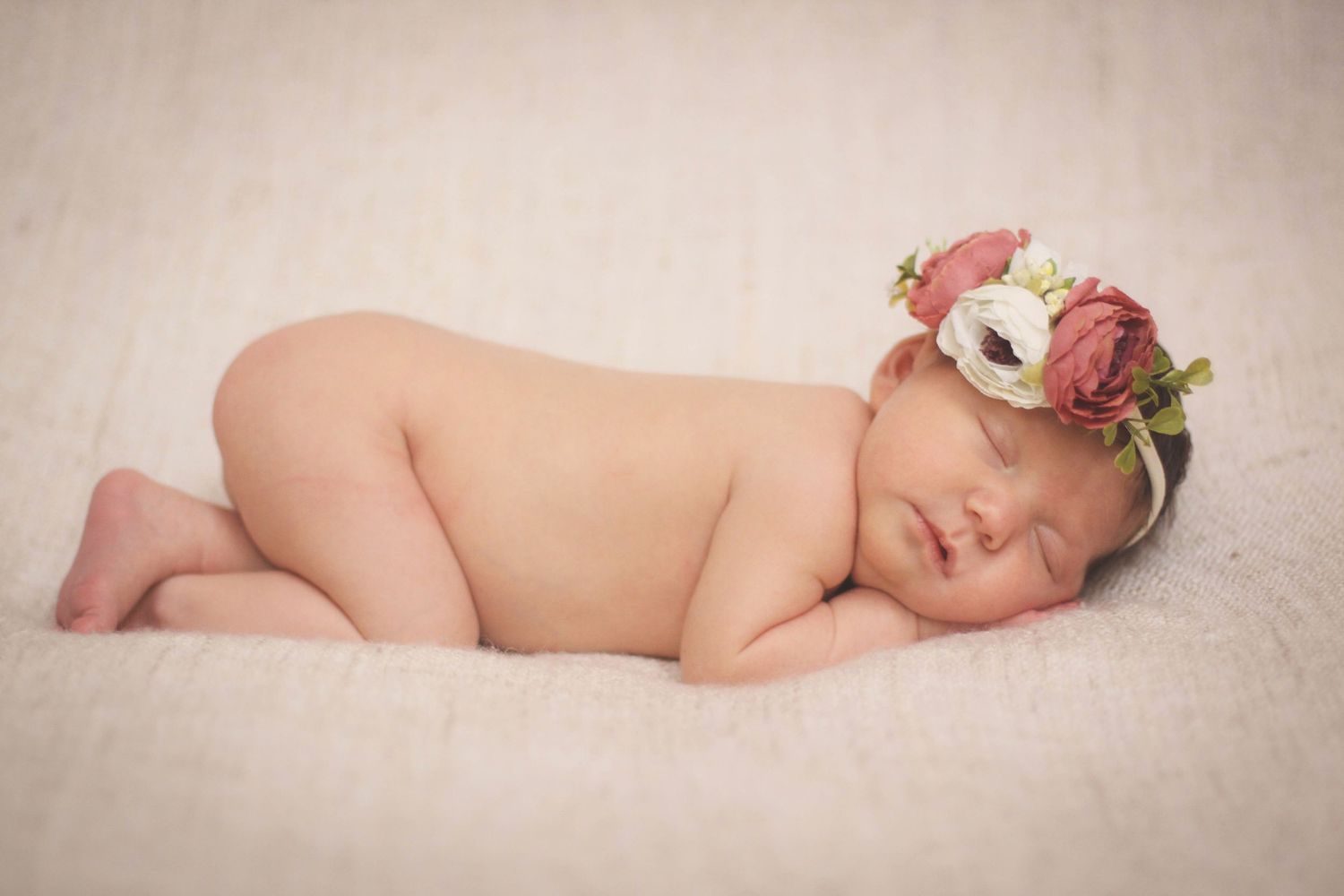 Sleeping newborn wearing delicate floral crown with pink and white flowers lies peacefully on soft beige fabric.