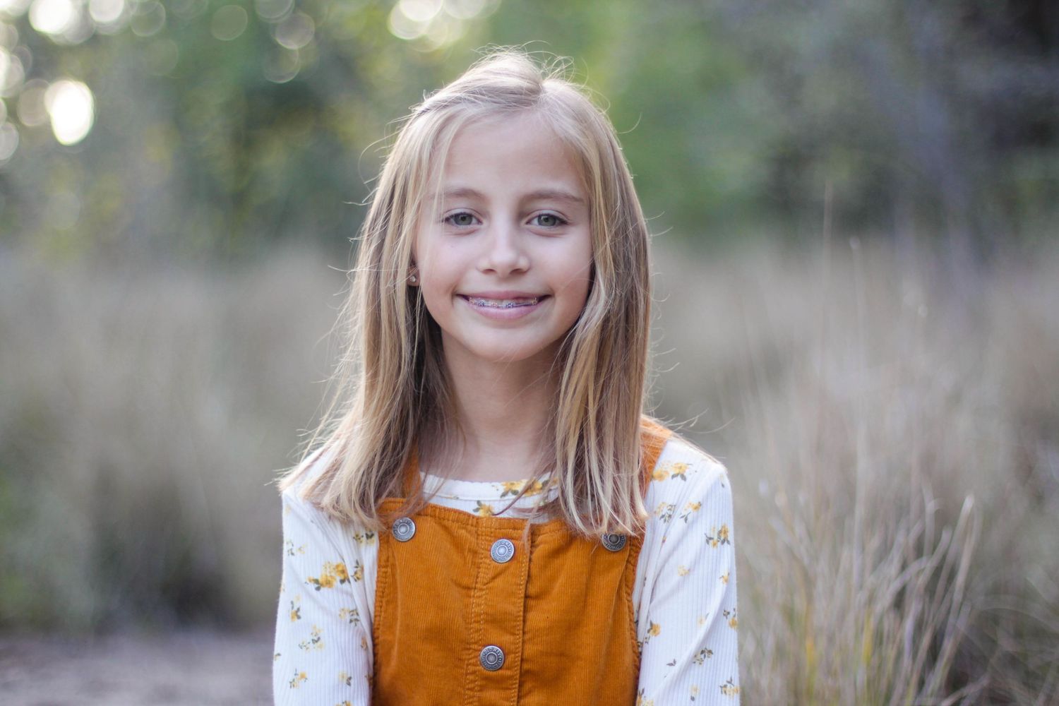Child in orange jumper dress photographed outdoors with soft bokeh background.