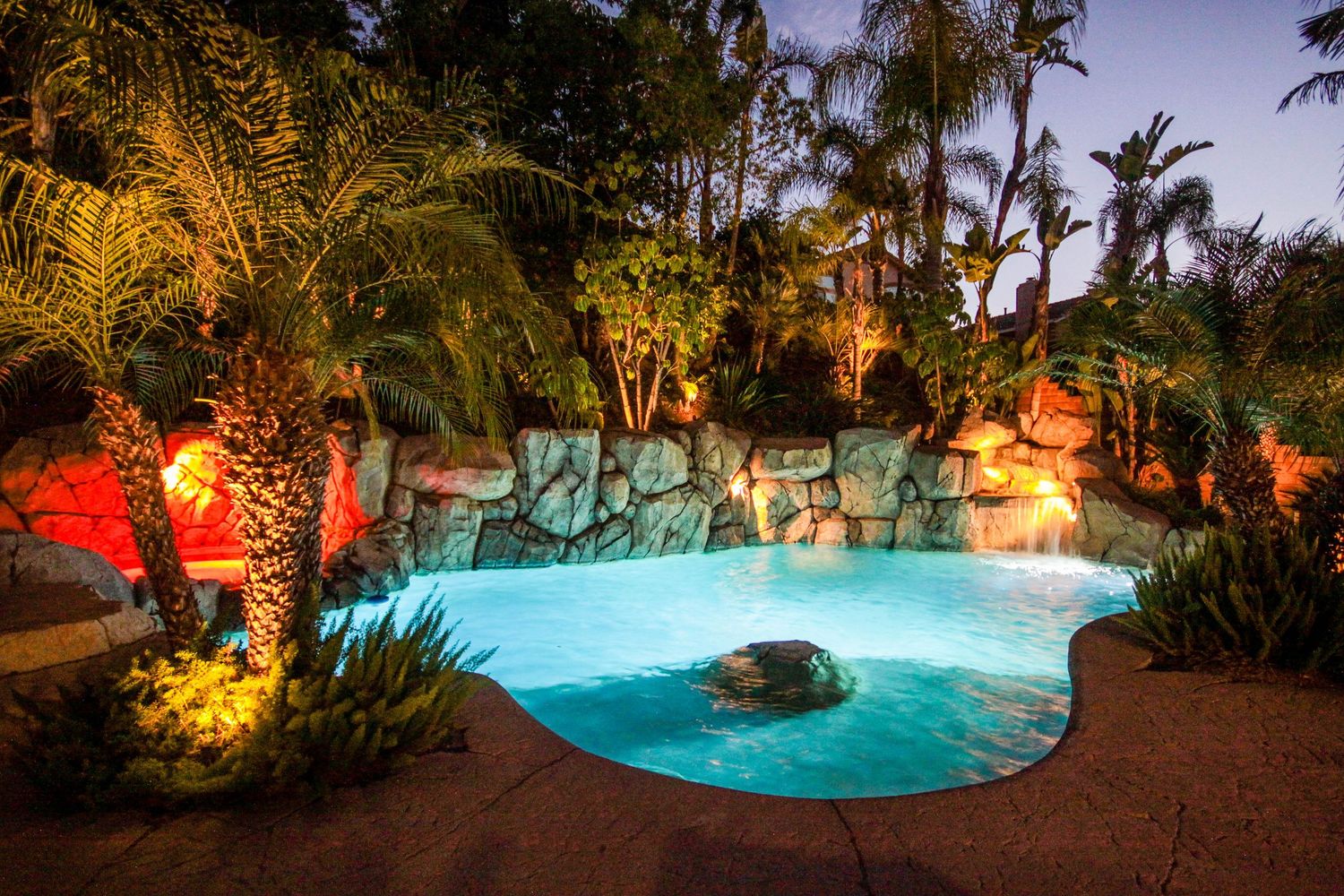 Curved swimming pool with rock features and palm trees lit up beautifully at night.