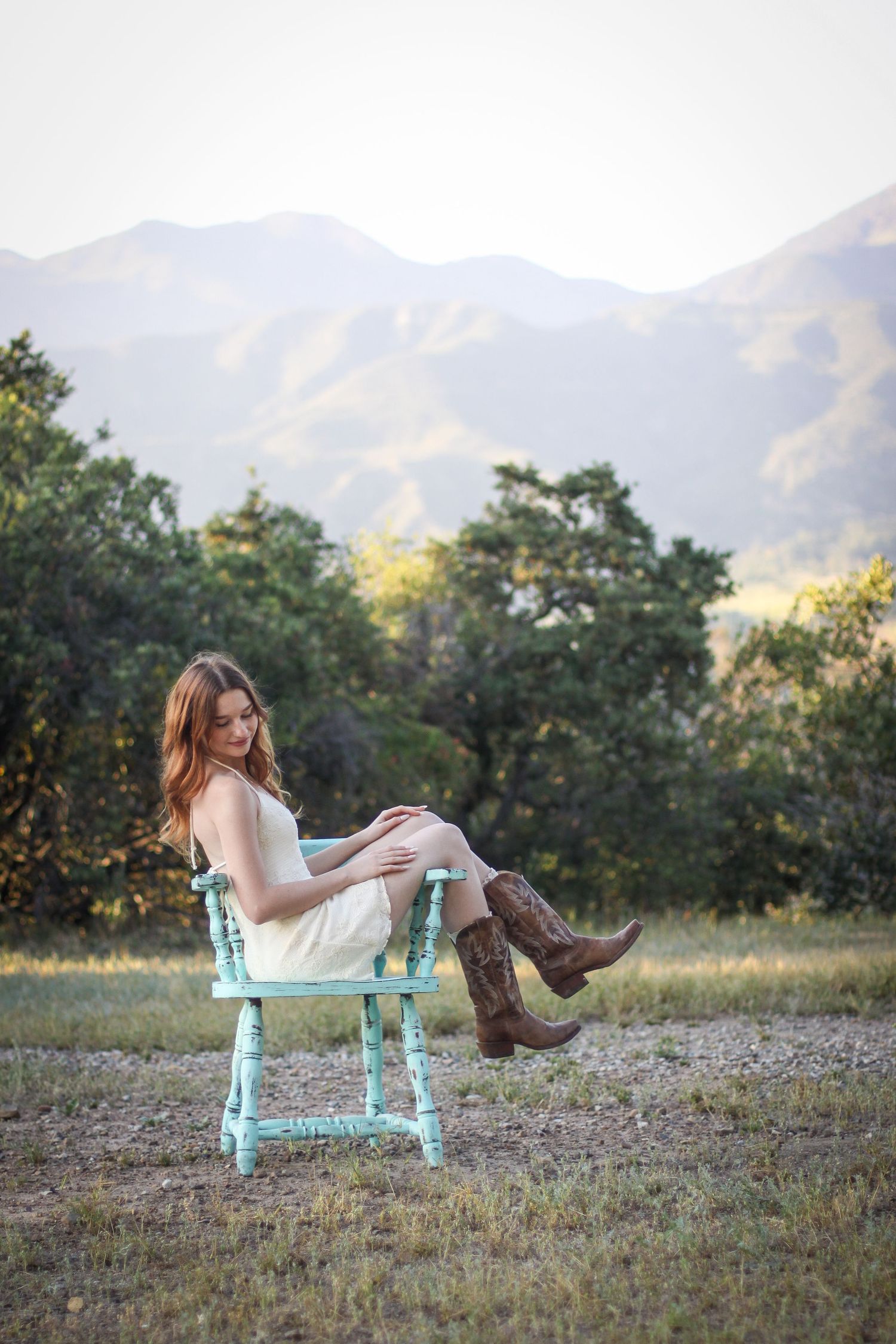 Woman in white dress and boots lounging on turquoise bench with mountains in background.