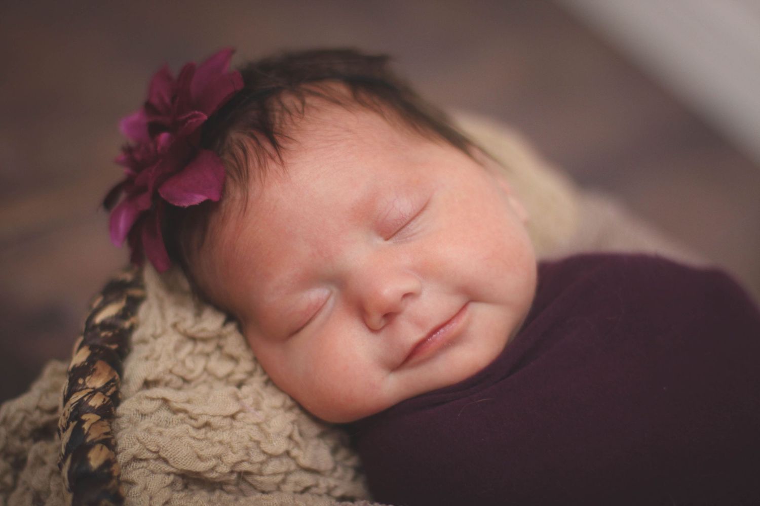 Newborn portrait with purple flower accessory against dark background.
