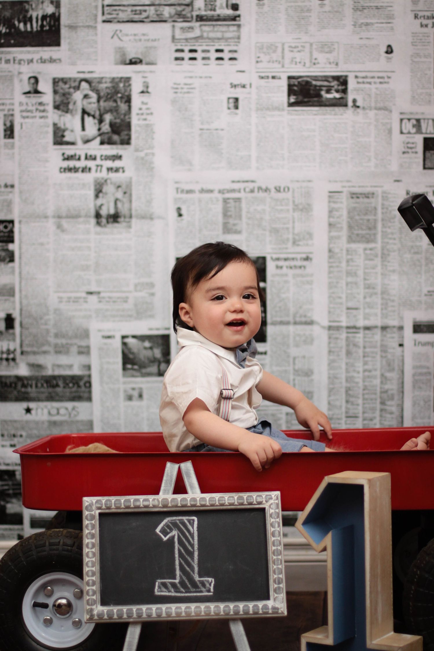 Baby in white outfit leaning over red table with newspaper background wall.
