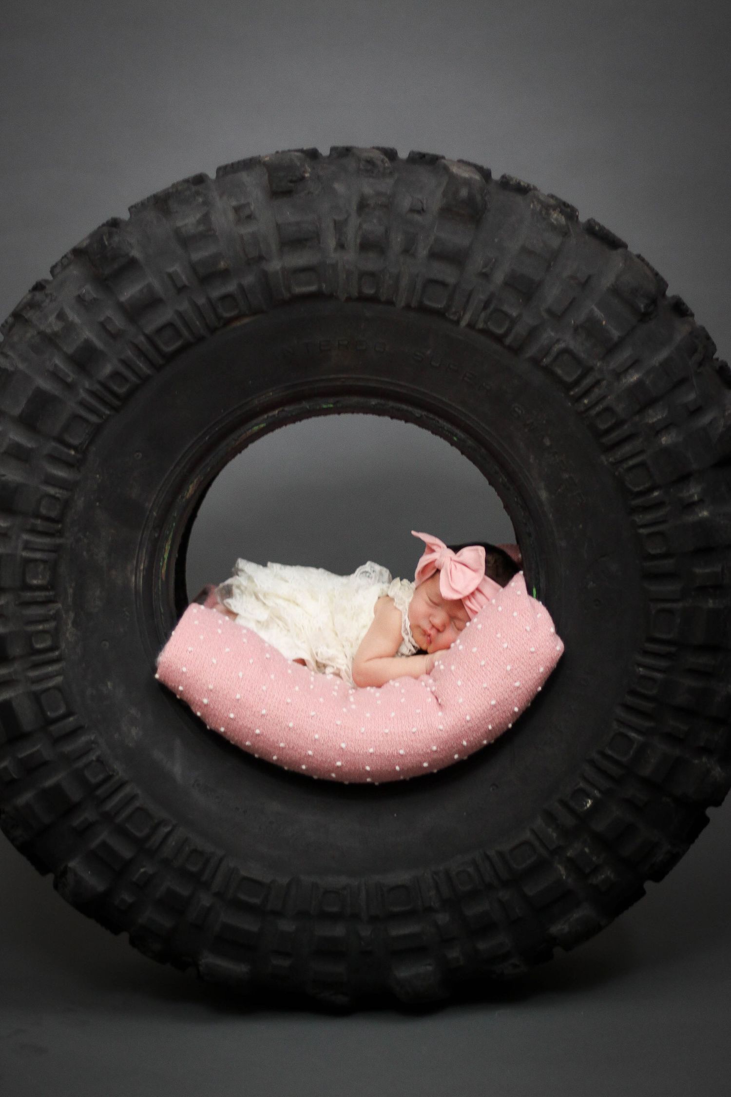 Sleeping newborn on pink cushion inside large black tire in dramatic lighting.