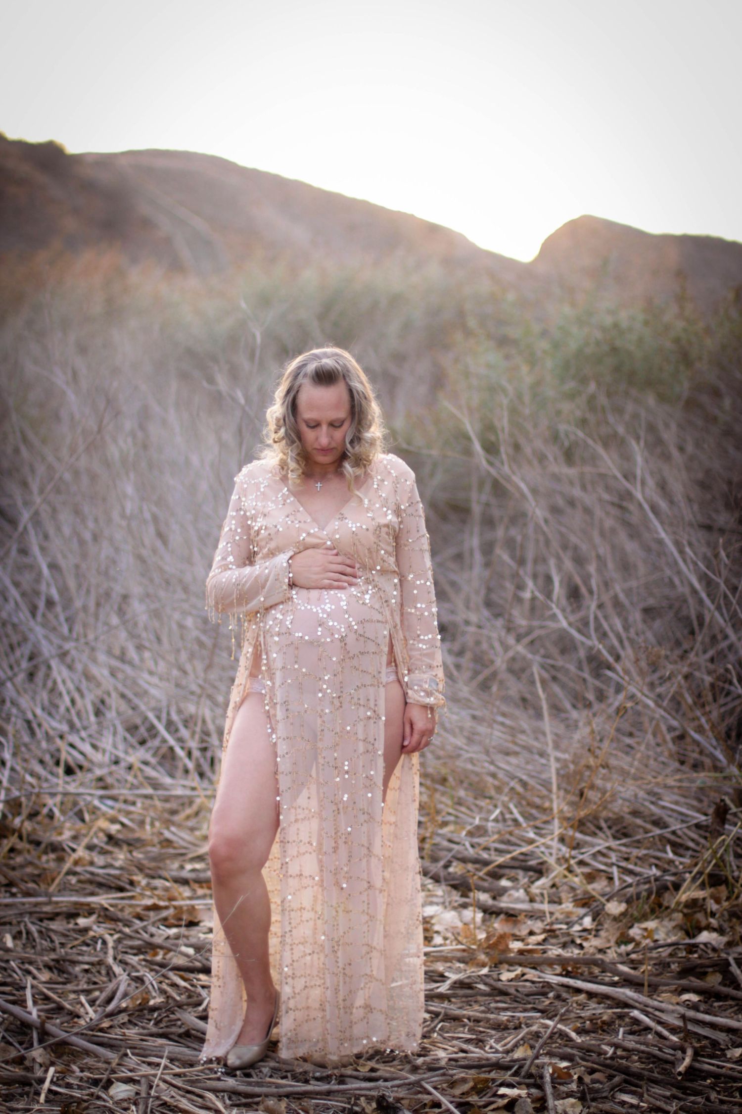 Ethereal maternity photo in sparkly blush gown against desert mountain landscape at sunset.