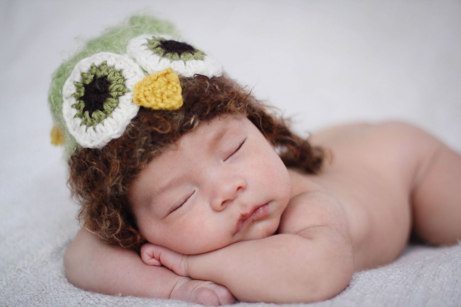 A sleeping baby wearing a delicate floral headpiece lies peacefully in a soft natural light setting.