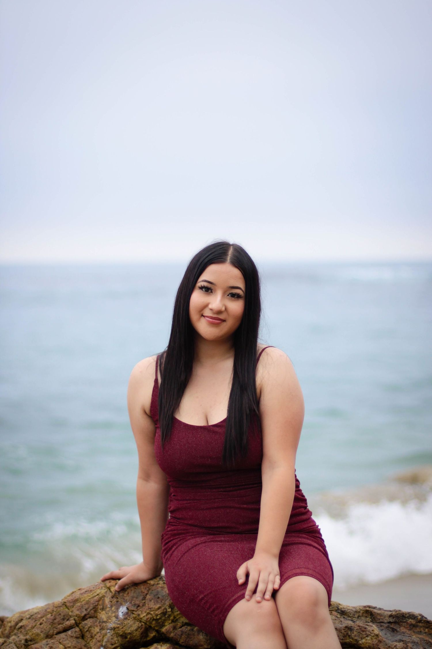Woman in burgundy dress sitting on coastal rocks by ocean.