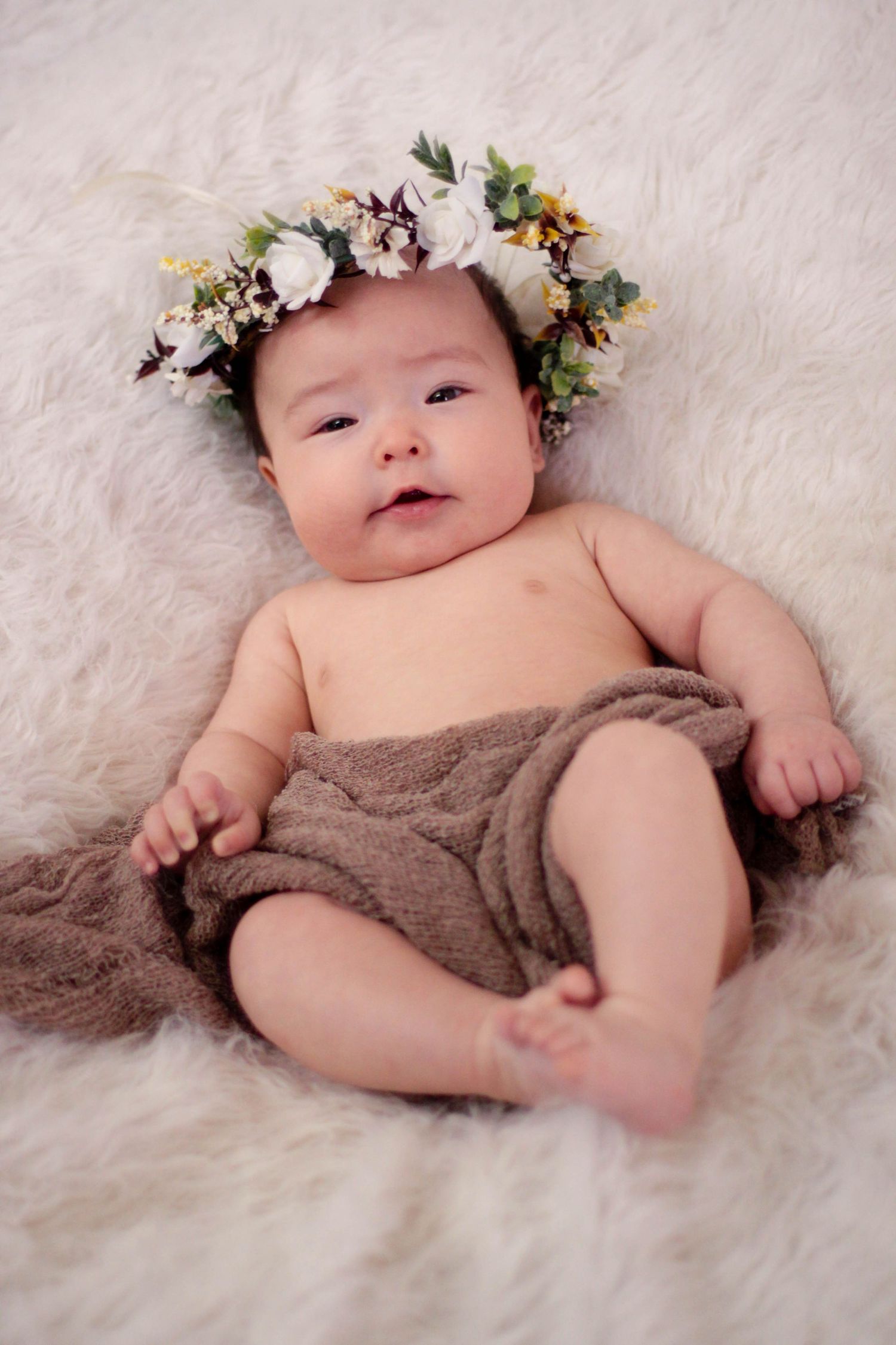 Baby lying on white fur rug wearing floral crown and wrapped in soft brown fabric.