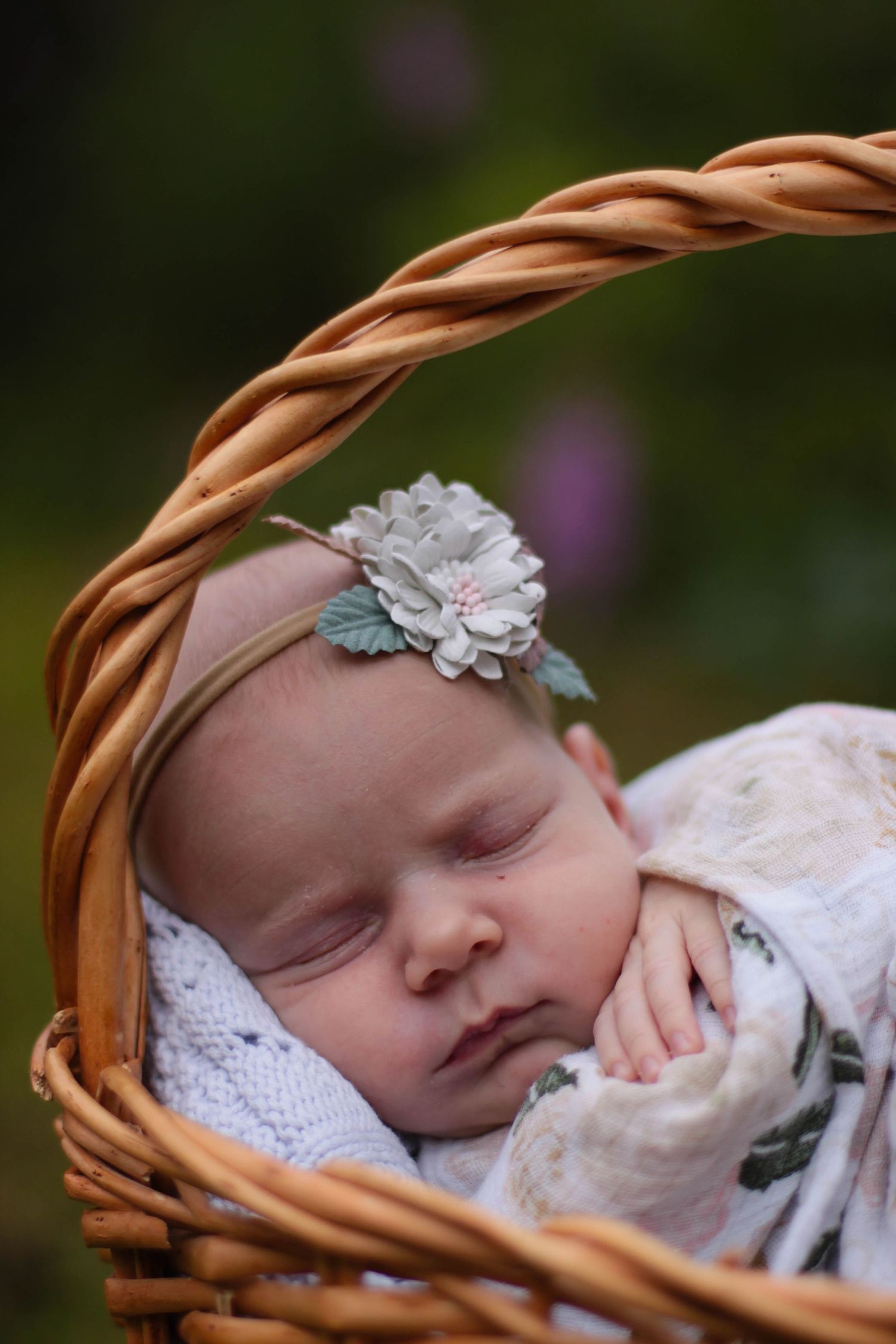 Newborn sleeping peacefully in wicker basket with floral headband and lace wrap.