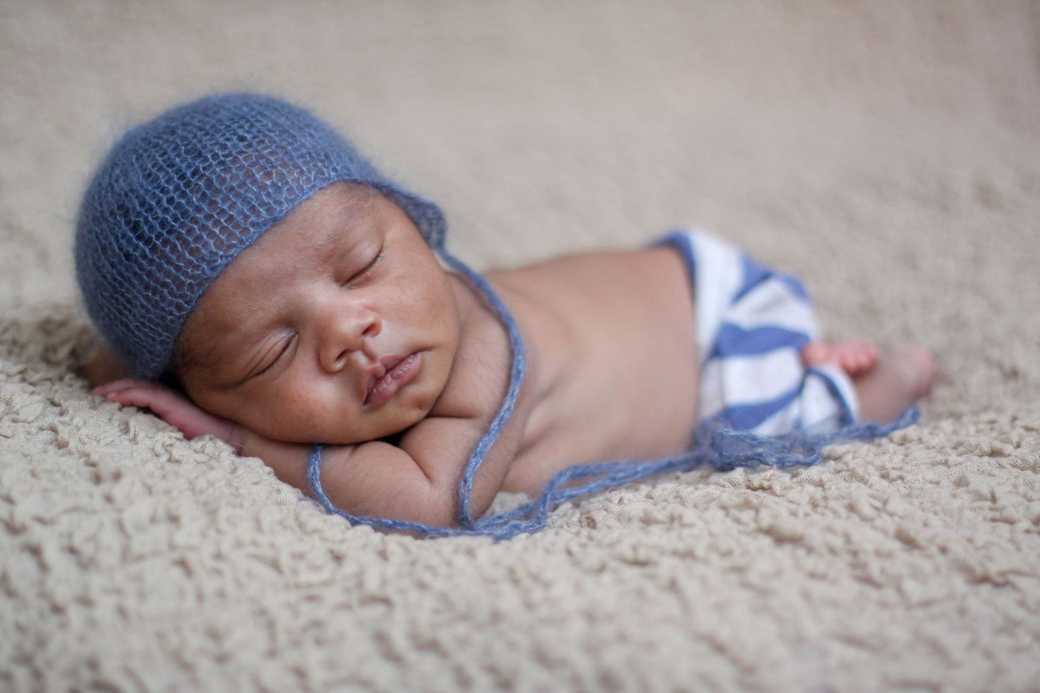 Sleeping newborn wearing a blue knit hat rests peacefully on soft gray carpet.