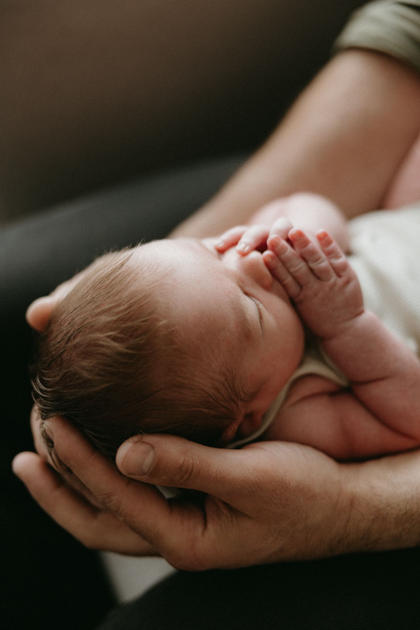 Newborn baby sleeping peacefully in parent's cupped hands, tiny fingers visible in a tender close-up portrait.