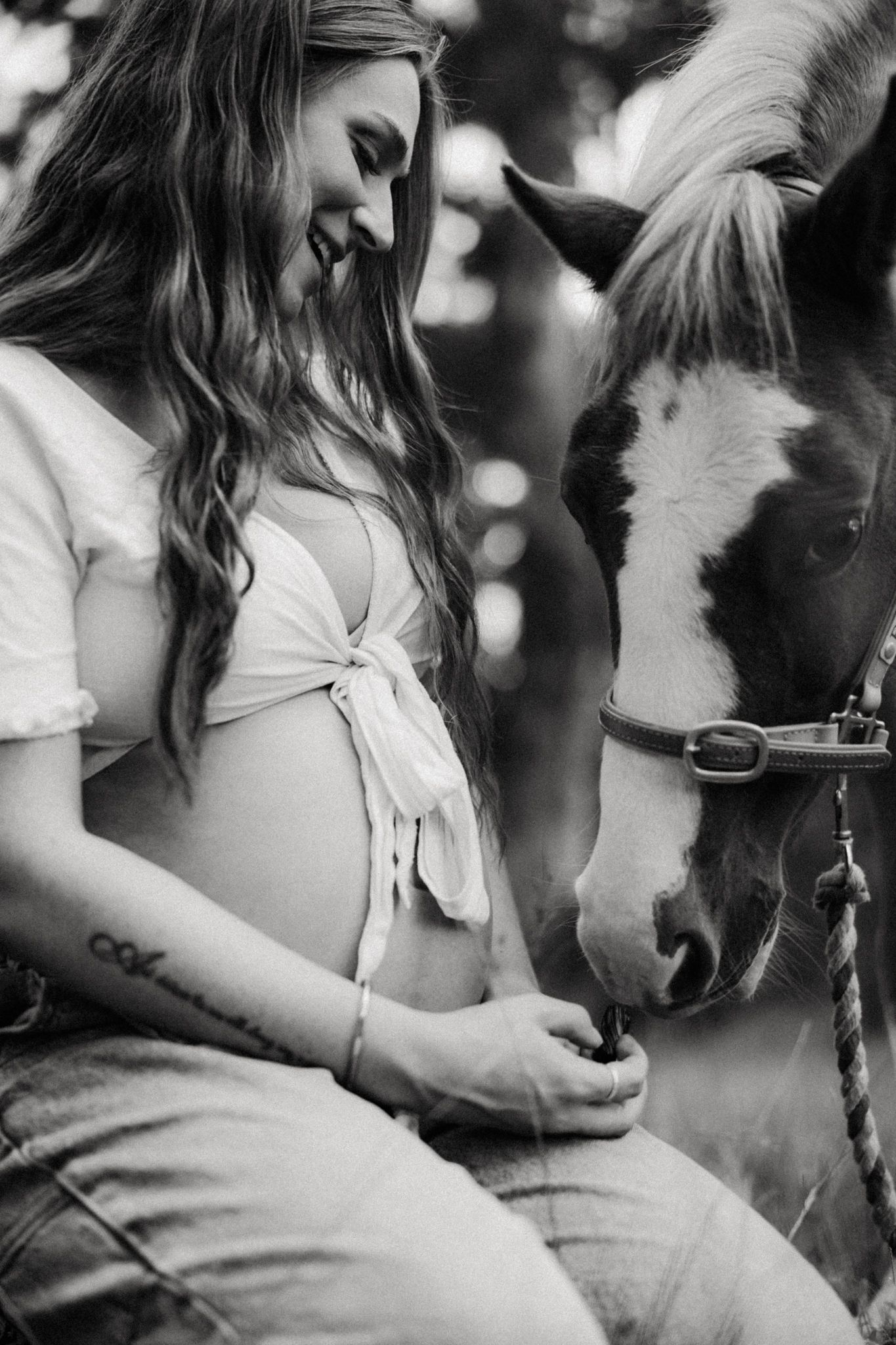 Black and white photo of smiling pregnant woman with exposed belly bonding with a horse outdoors.