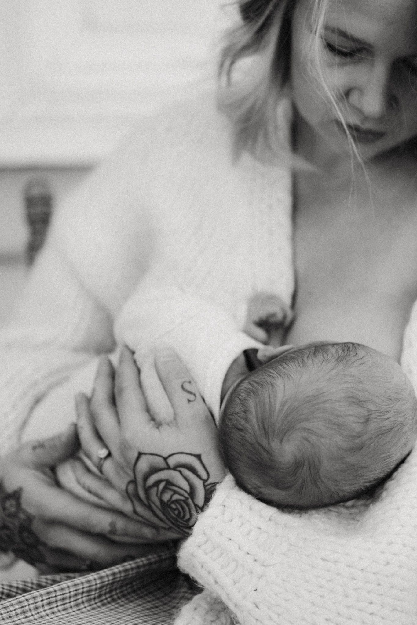 Black and white photo of tattooed mother breastfeeding her newborn baby, wrapped in a white knit blanket.