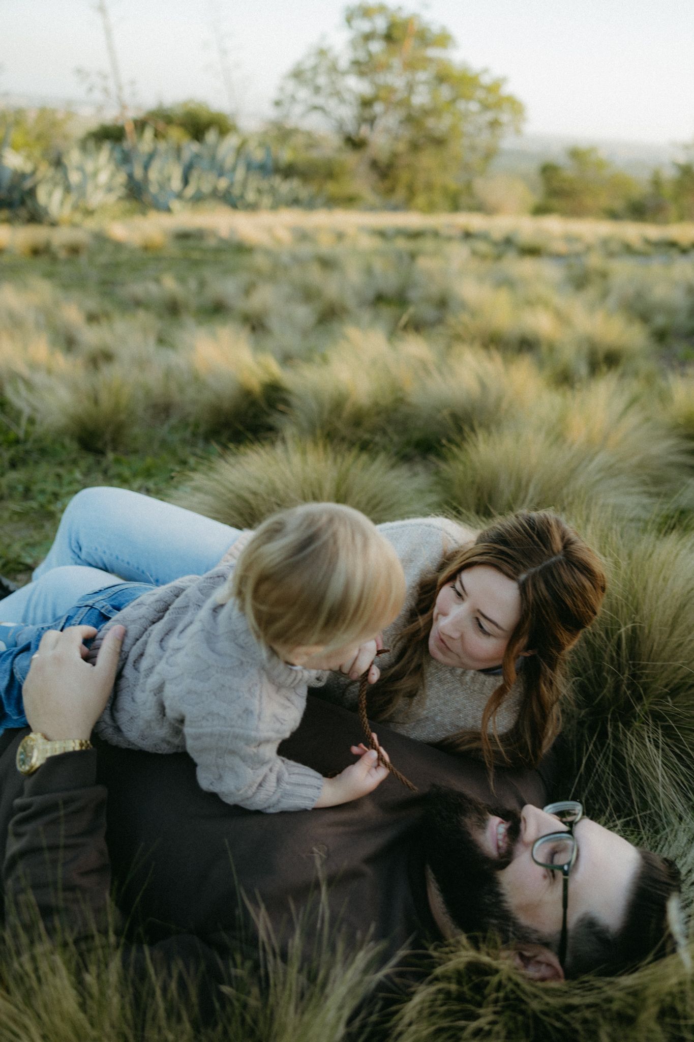 A young family relaxing together in a grassy field, with a toddler playing on parents lying in the grass.
