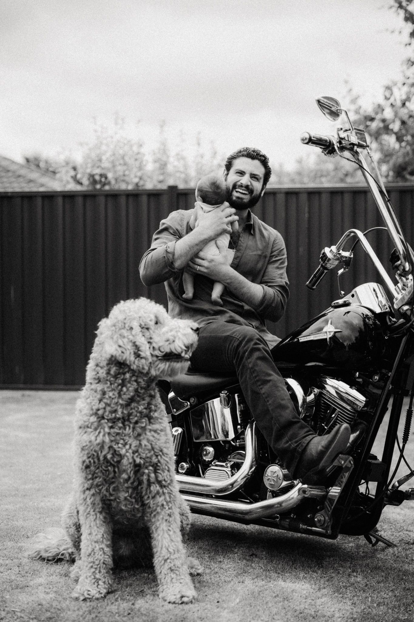 Bearded man sitting on a classic motorcycle holding a baby, with a fluffy poodle dog in the foreground, black and white photo.