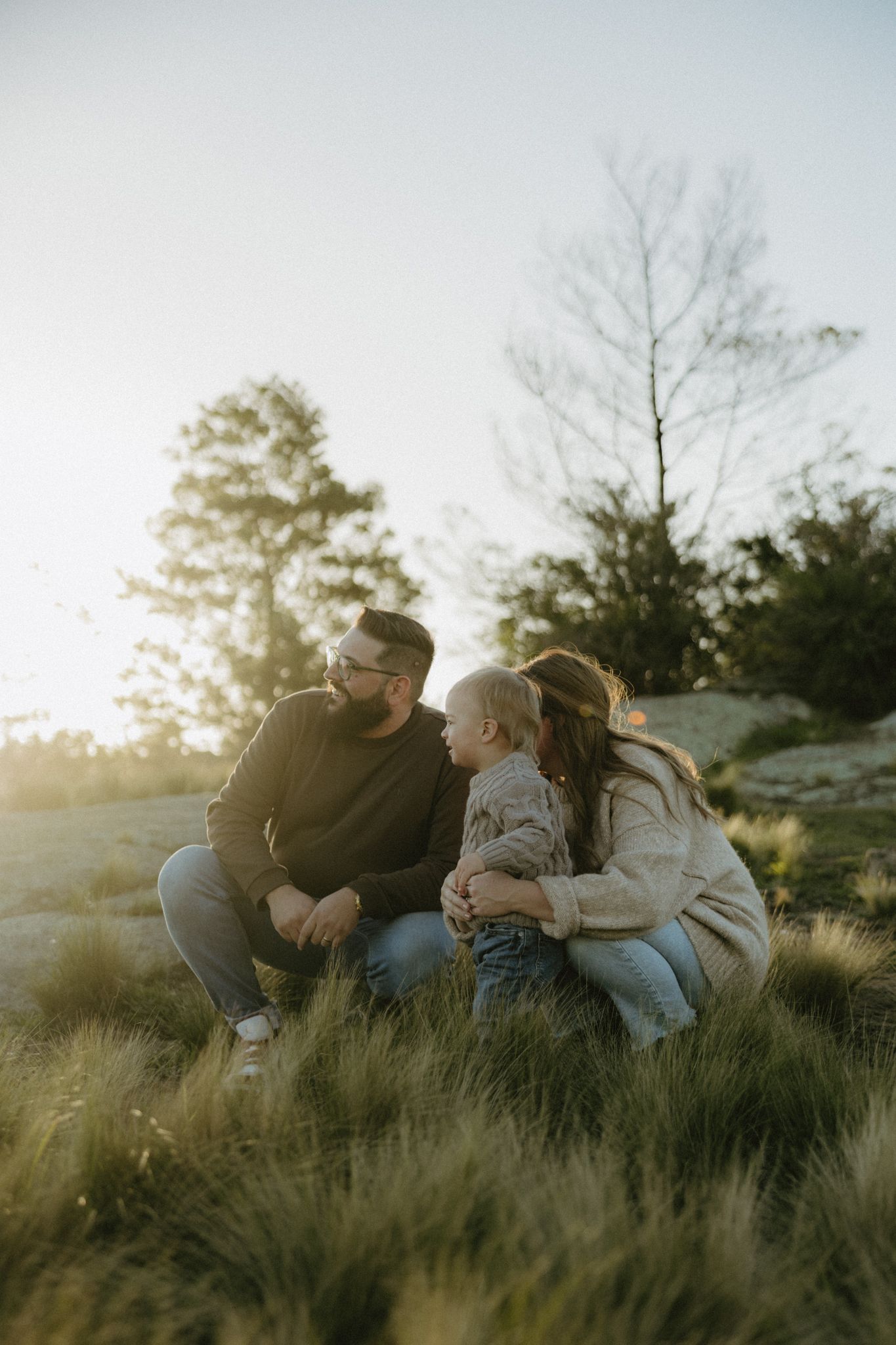 Family of three sharing a tender moment outdoors at golden hour, with trees and soft sunlight in the background.