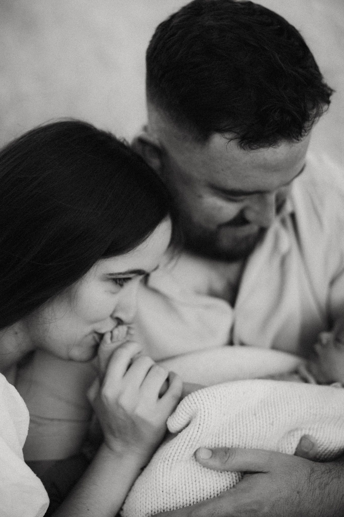 Black and white photo of parents tenderly kissing their newborn baby wrapped in a soft knit blanket.