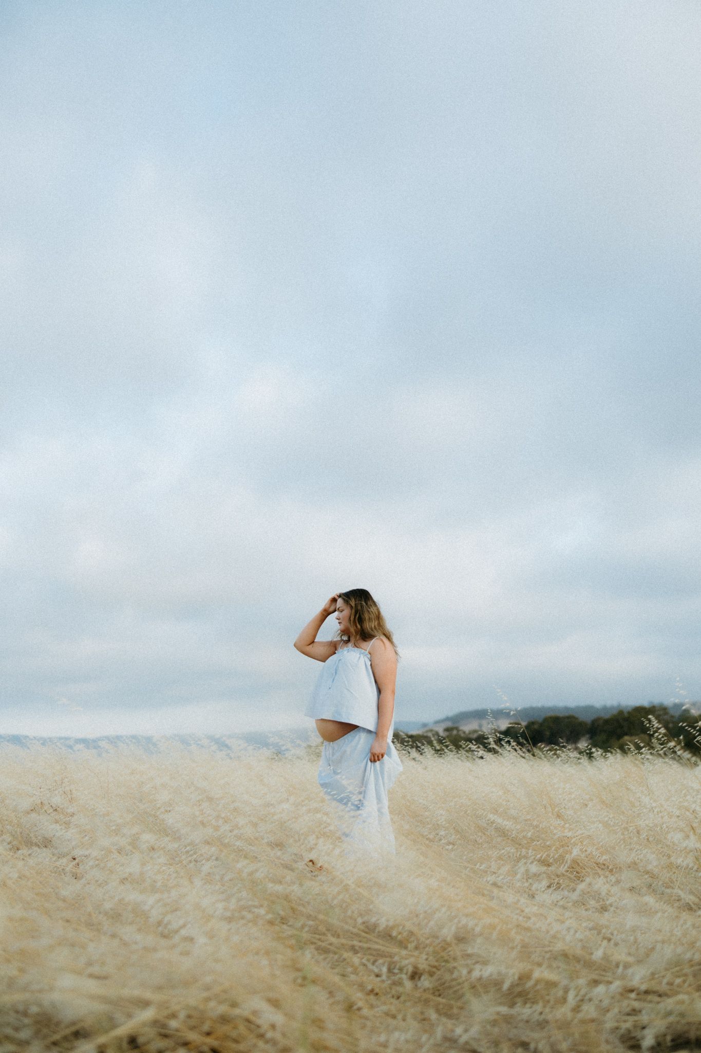 Pregnant woman in white crop top and skirt standing in golden wheat field under cloudy sky.
