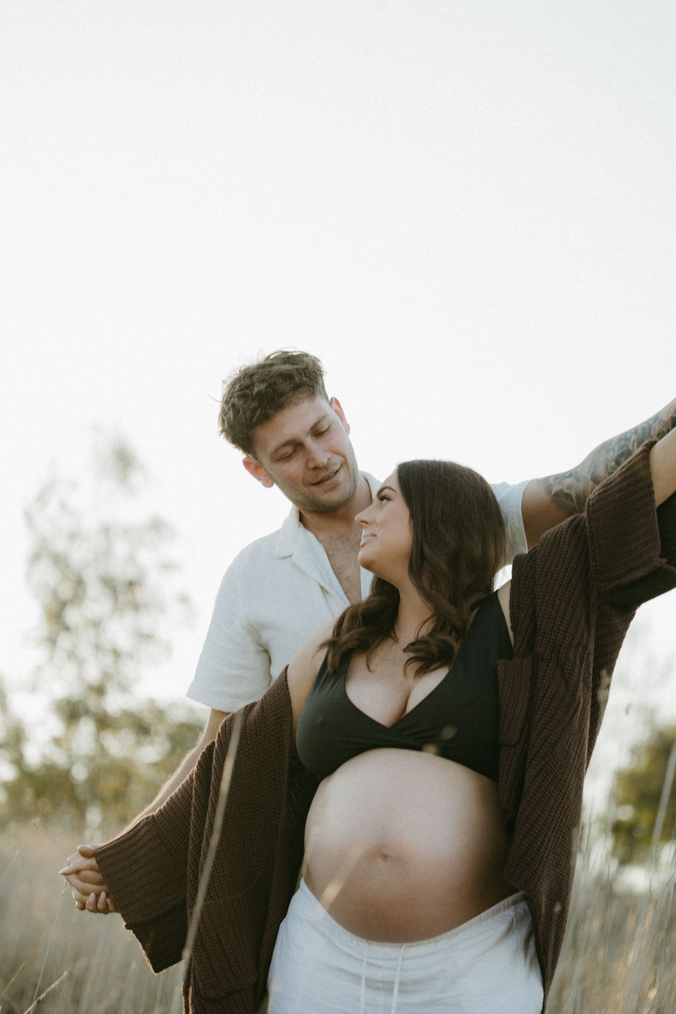 Couple shares tender moment outdoors, pregnant woman in black crop top and cardigan smiling up at partner in sunlit field.