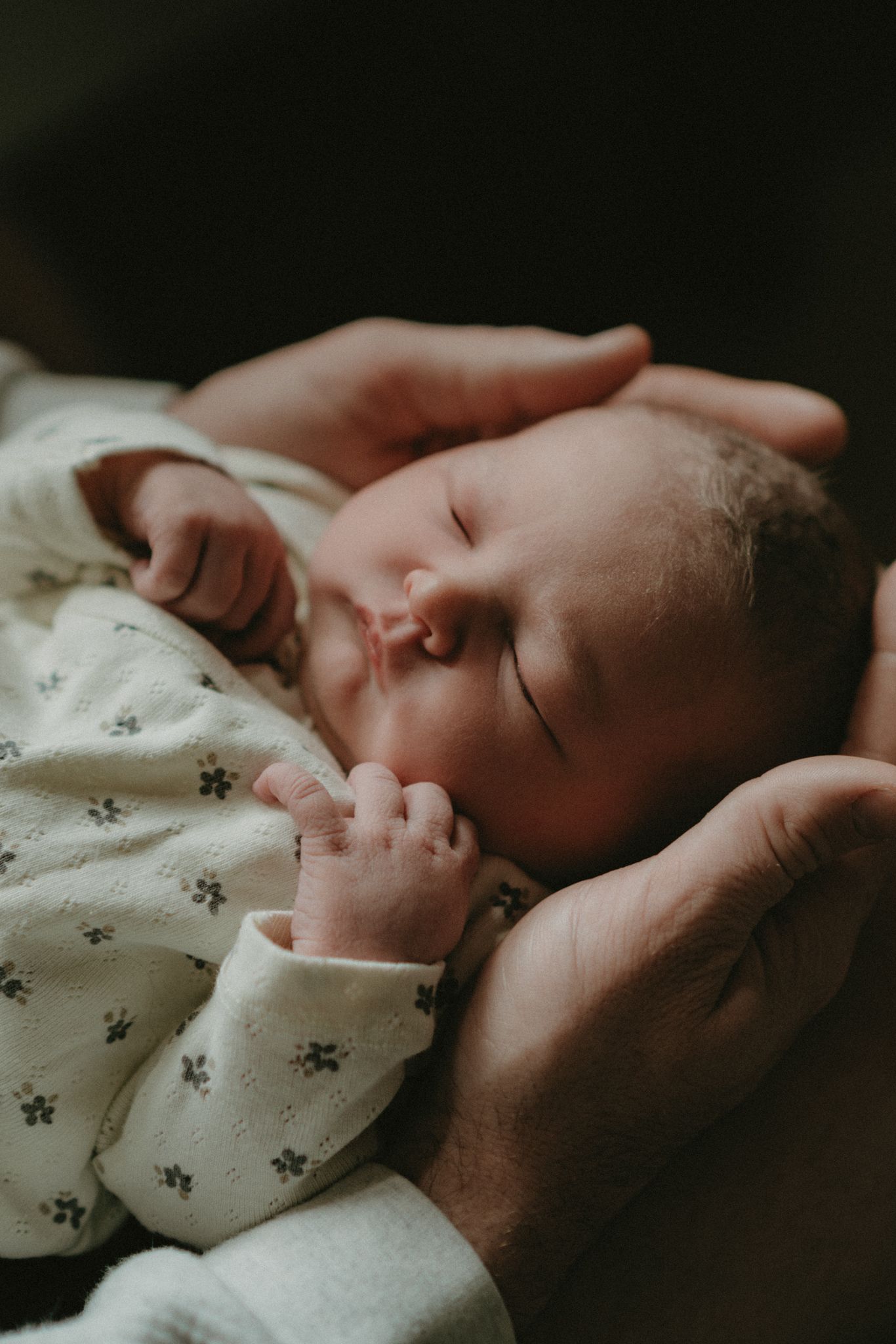 A peaceful newborn baby sleeping in a parent's gentle hands, wearing a white floral onesie in soft natural light.