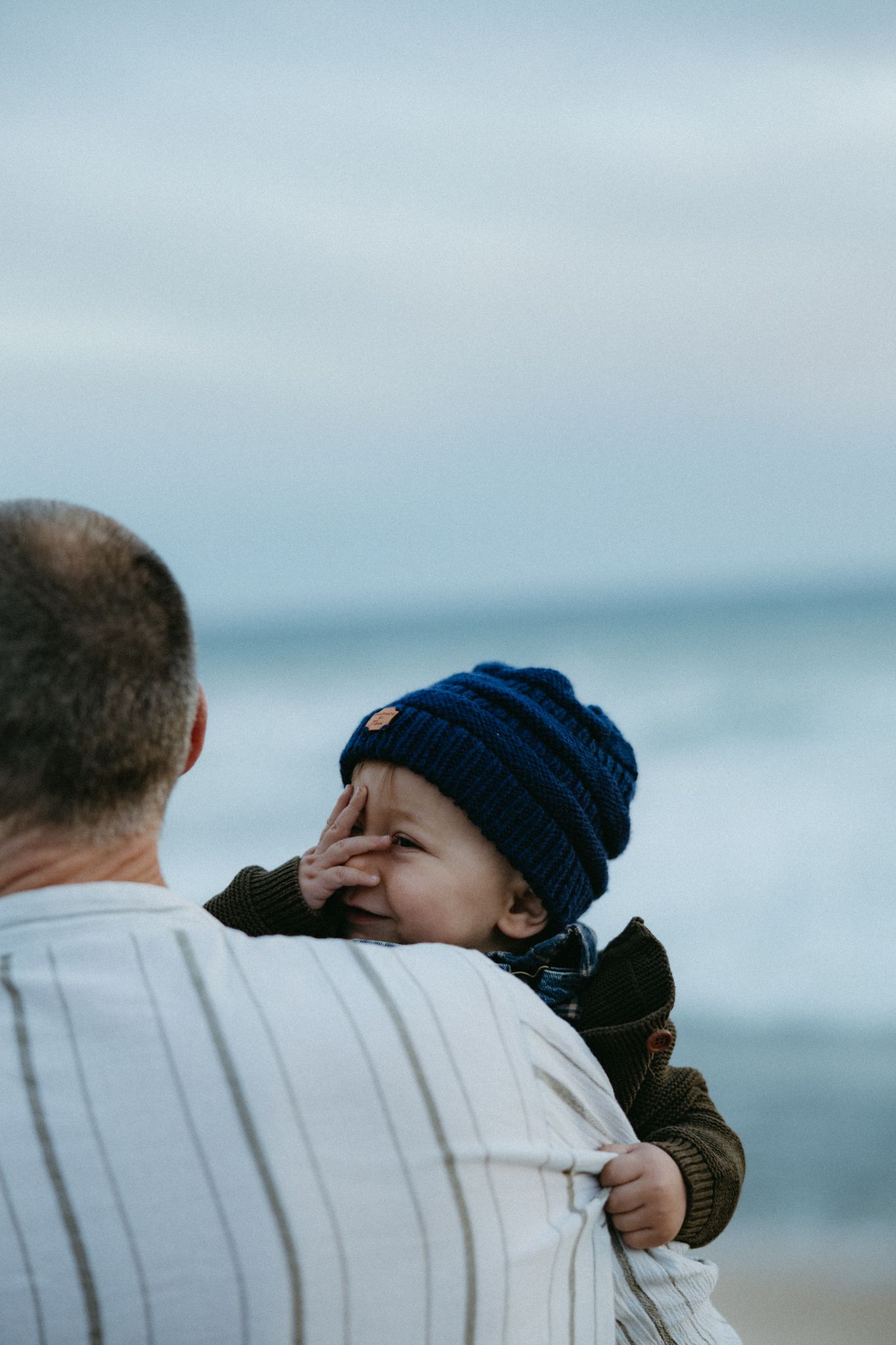 An adult holds a baby wearing a blue knit beanie at the beach, with ocean waves in the background.