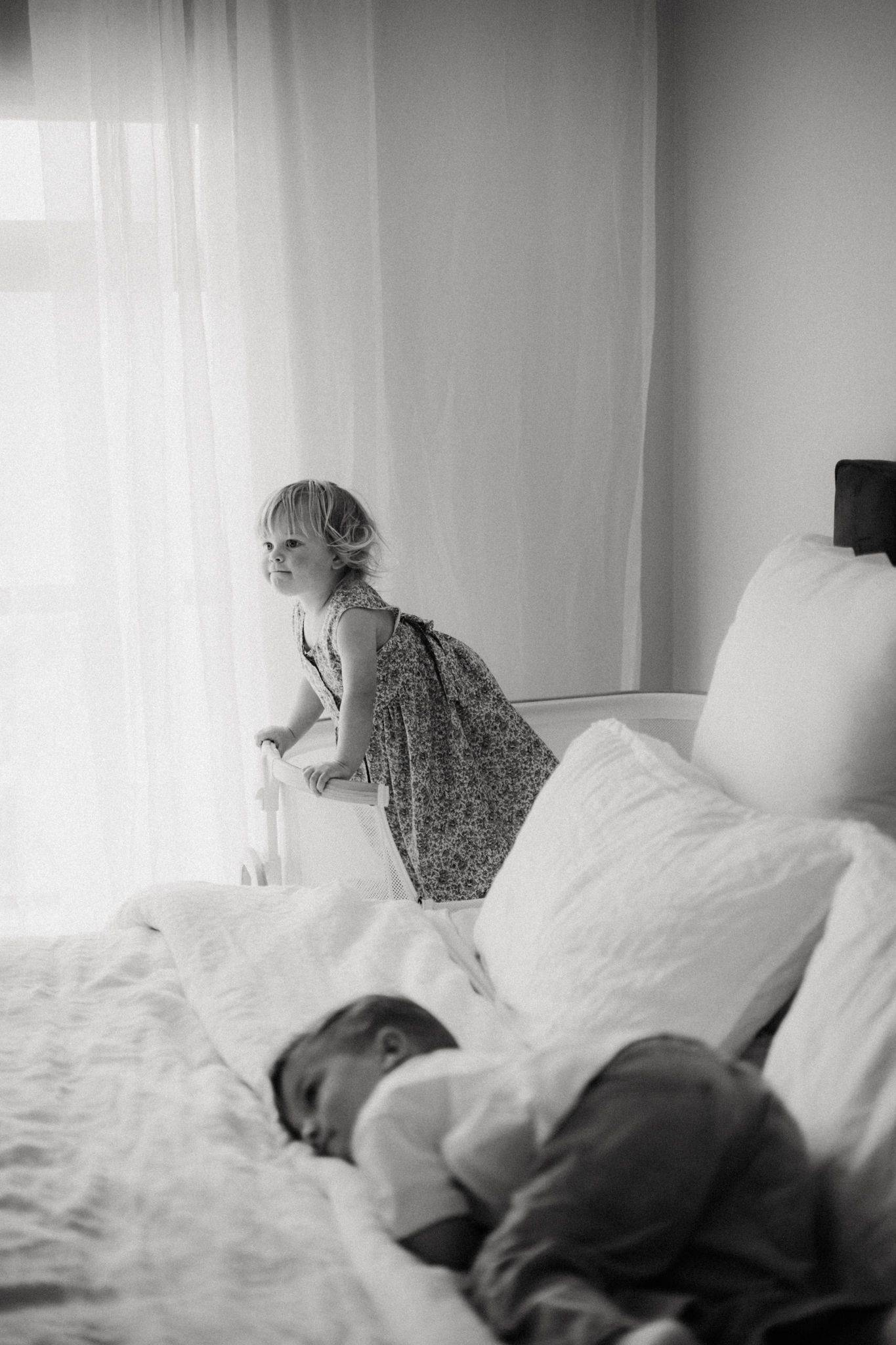Black and white photo of a toddler standing by a bright window beside a sleeping newborn on a white bed.