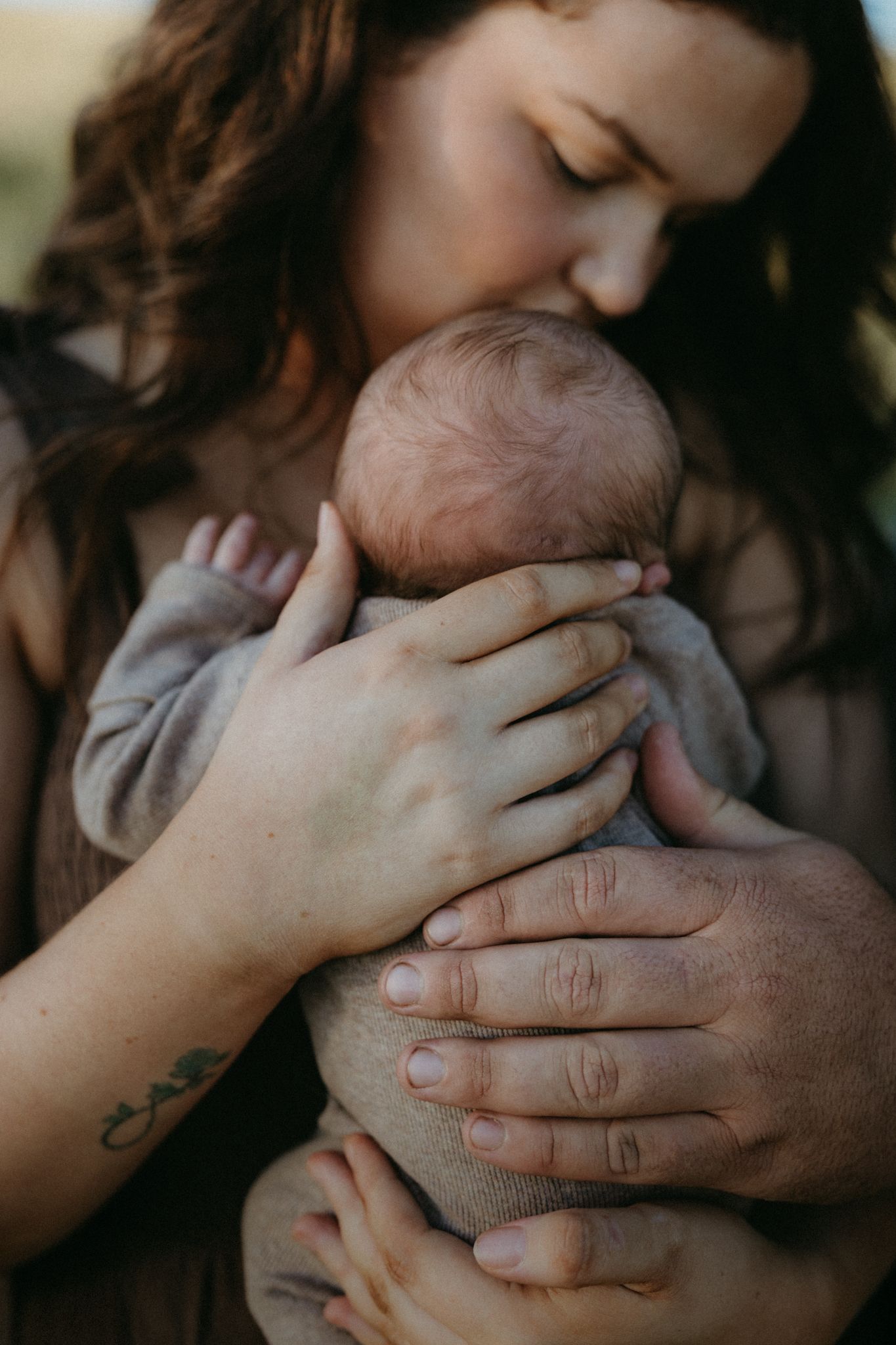 A mother tenderly kisses her newborn baby while a partner's hands support the infant in a warm outdoor embrace.
