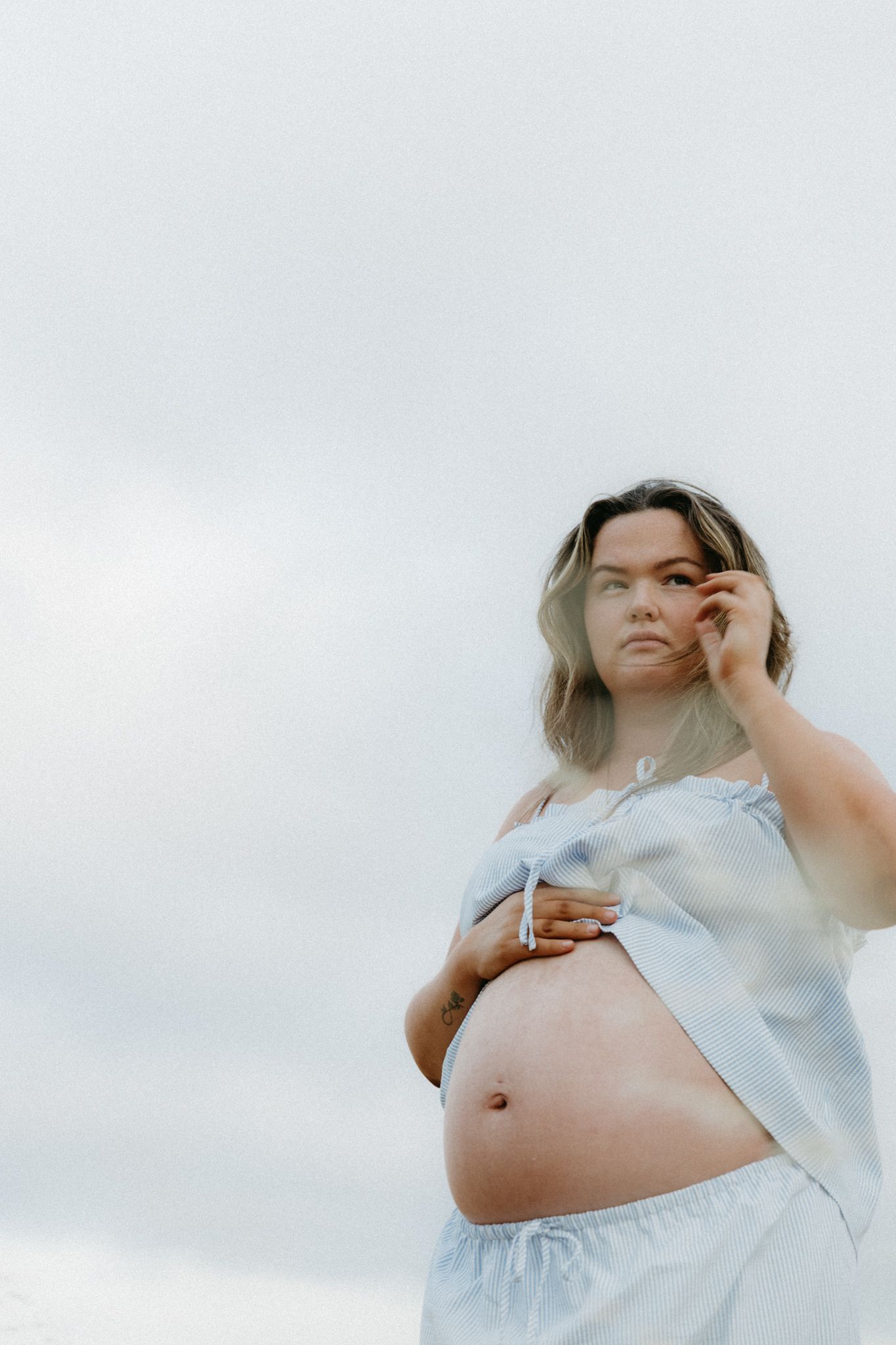 Pregnant woman in blue crop top exposing baby bump, standing outdoors against cloudy sky.
