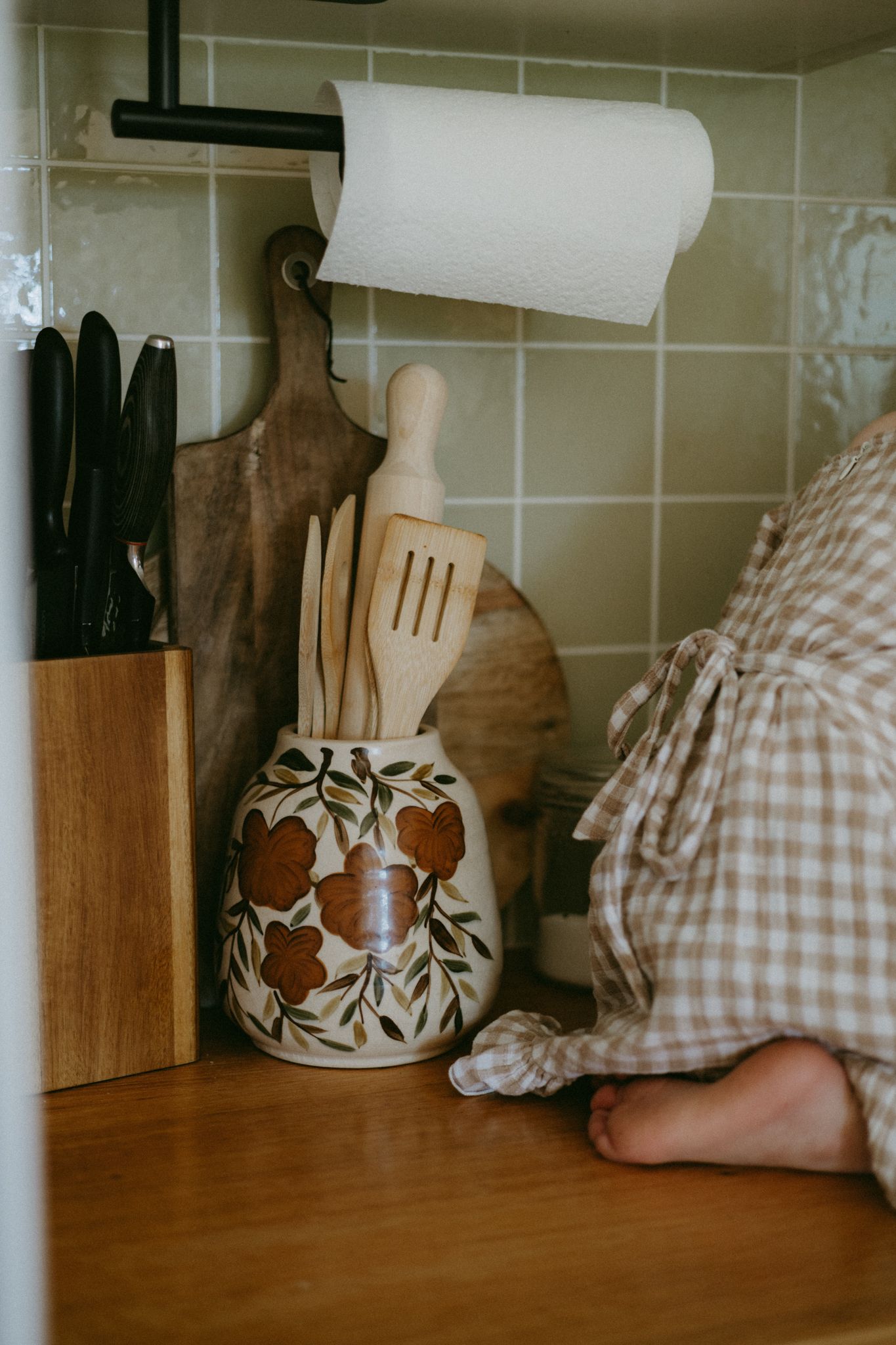 Rustic kitchen counter with wooden utensils in floral ceramic crock, knife block, and paper towel holder.
