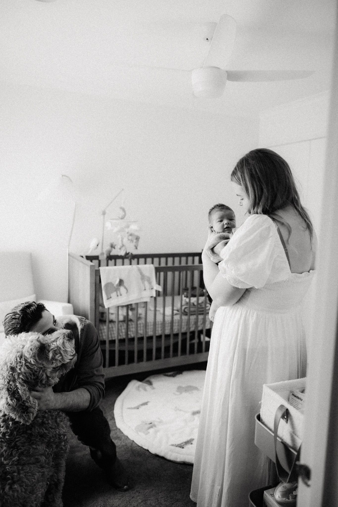 Black and white photo of a mother in white dress holding newborn baby in nursery while man pets fluffy dog nearby.