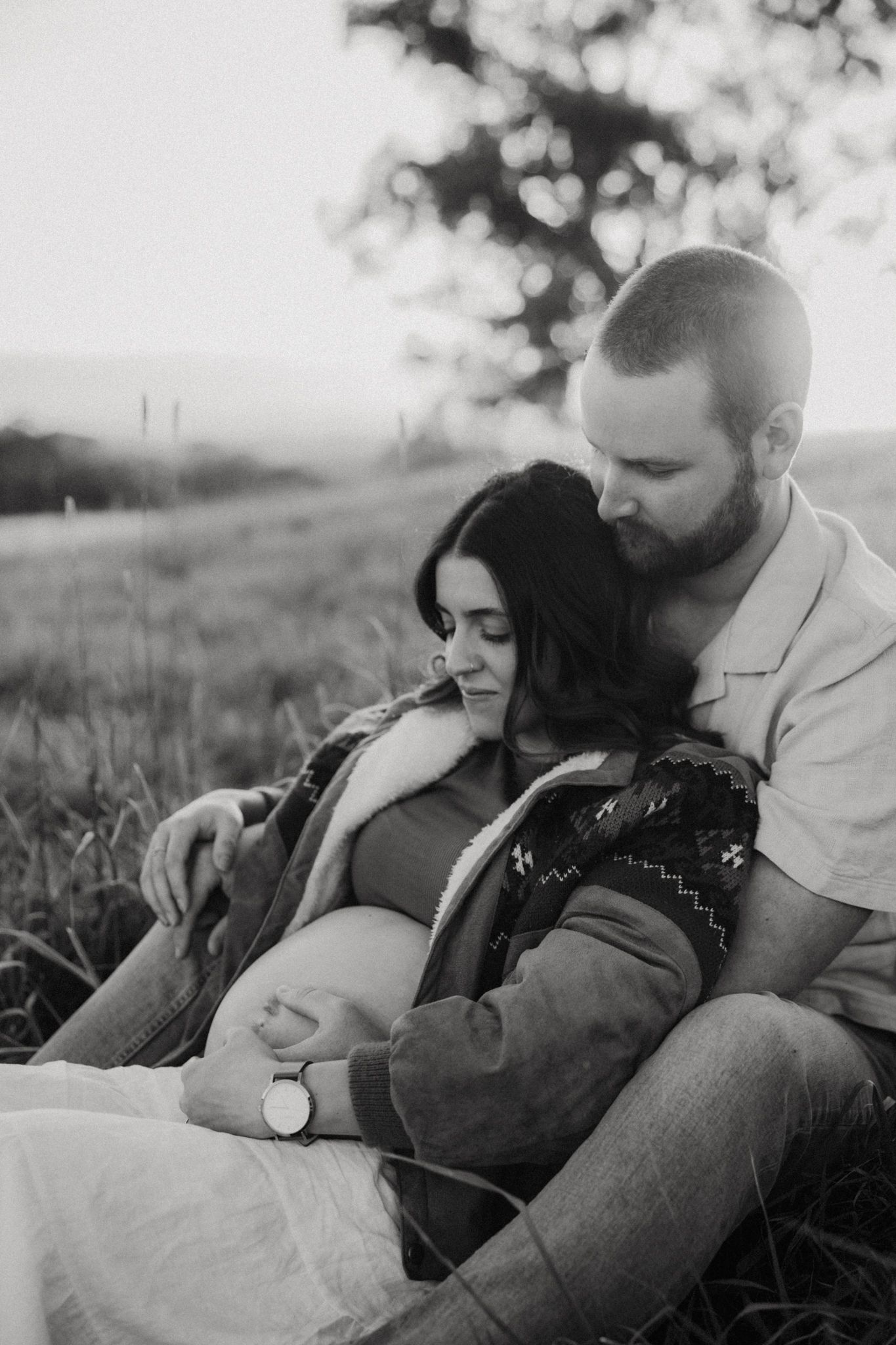 Black and white maternity photo of a couple sitting in a field, man embracing pregnant woman at sunset.
