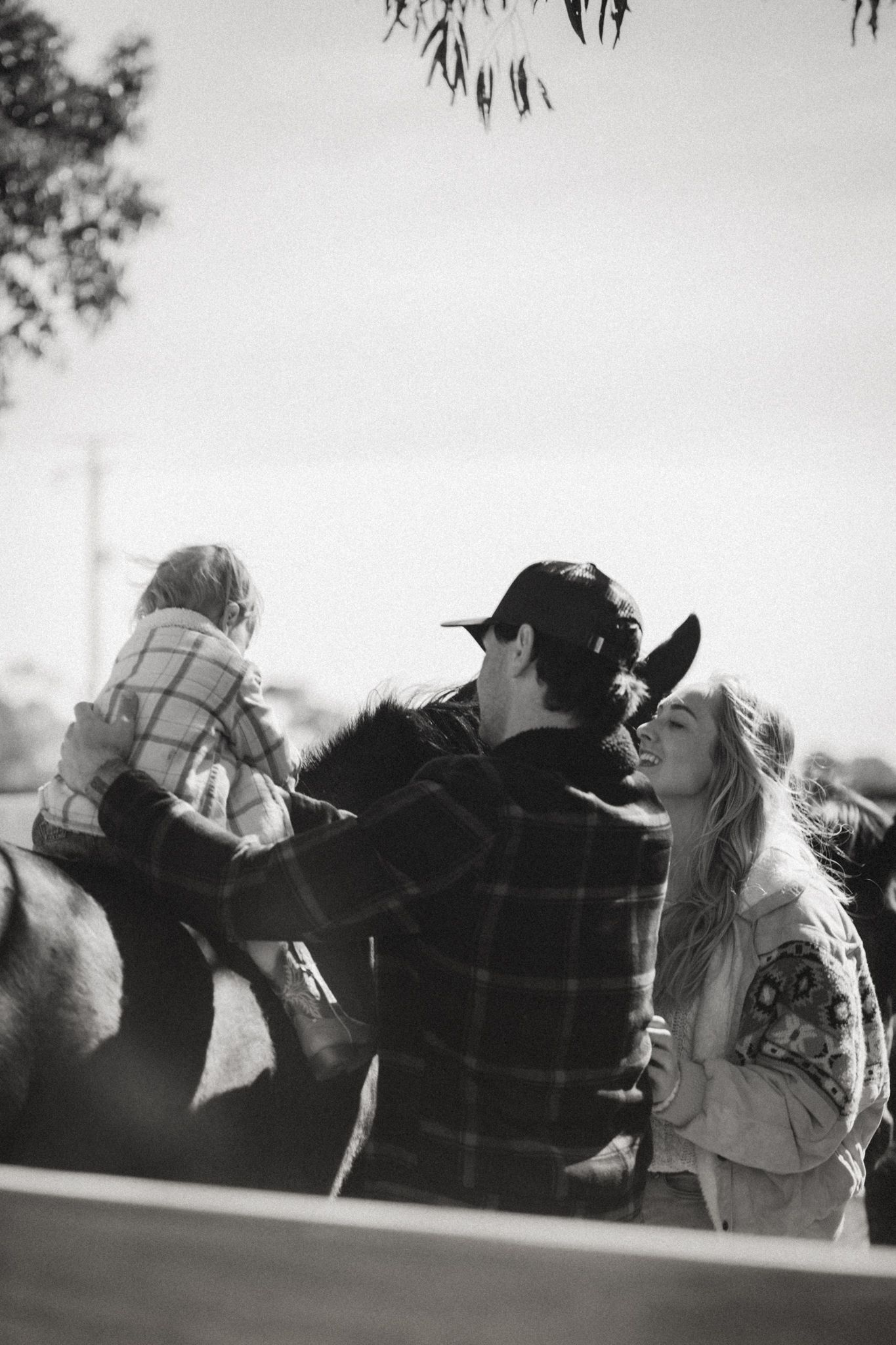 Black and white photo of a couple sharing a tender moment while a child sits on a horse at a ranch.