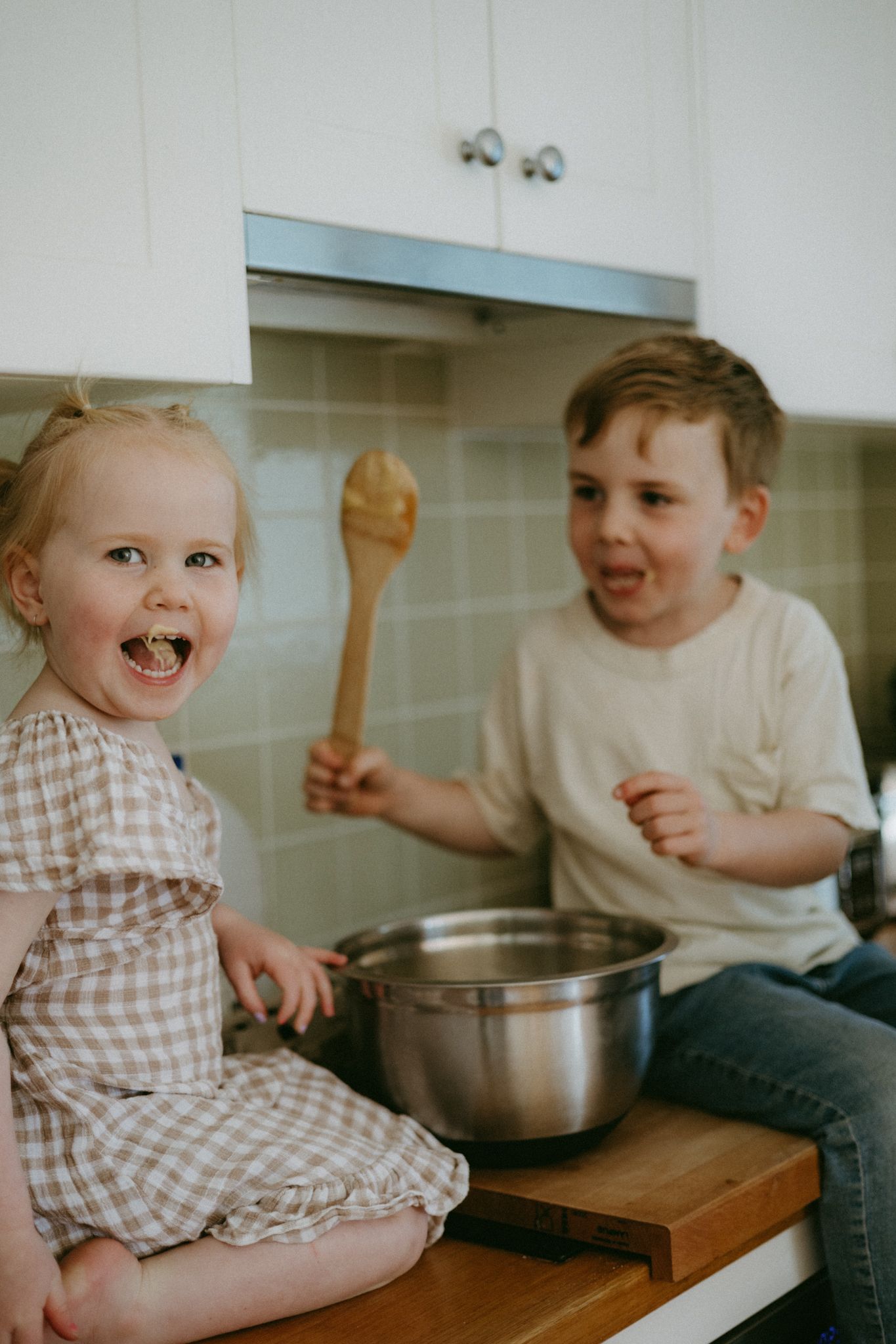 Two happy young children sitting on a kitchen counter with a mixing bowl and wooden spoon.
