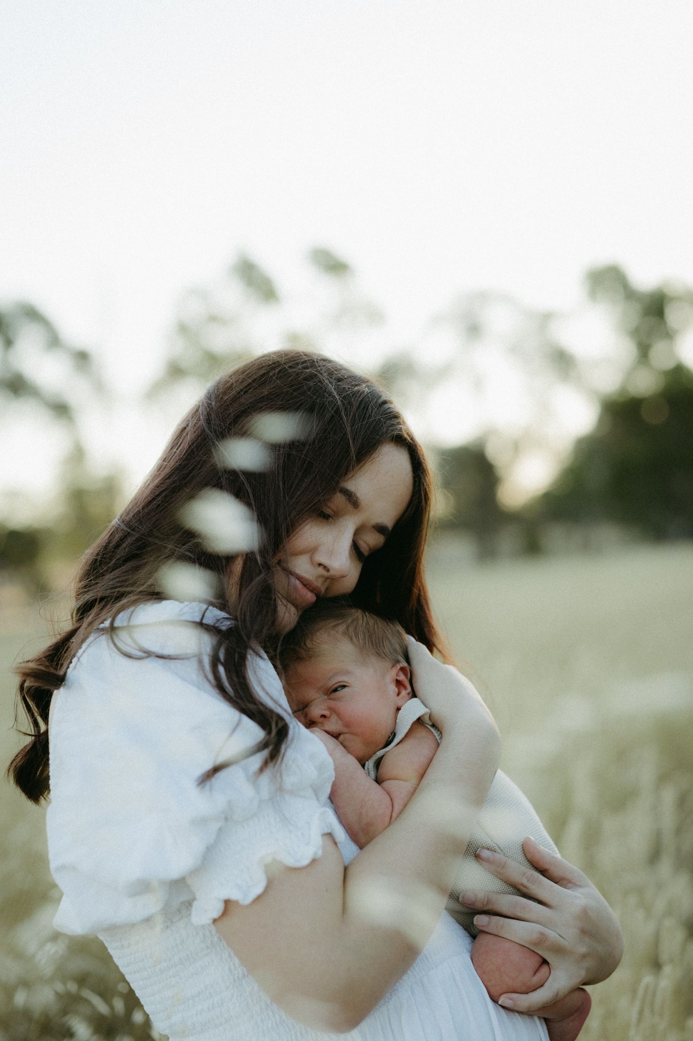 A mother in a white dress tenderly holds her newborn baby in a sunlit field with soft bokeh background.