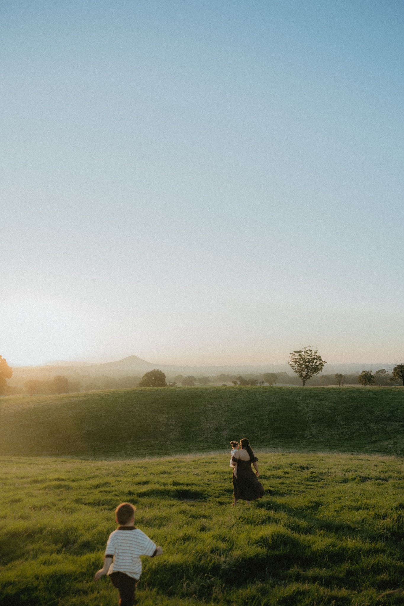 Child running toward a woman in a sunlit green field at golden hour with rolling hills and a lone tree in the background.