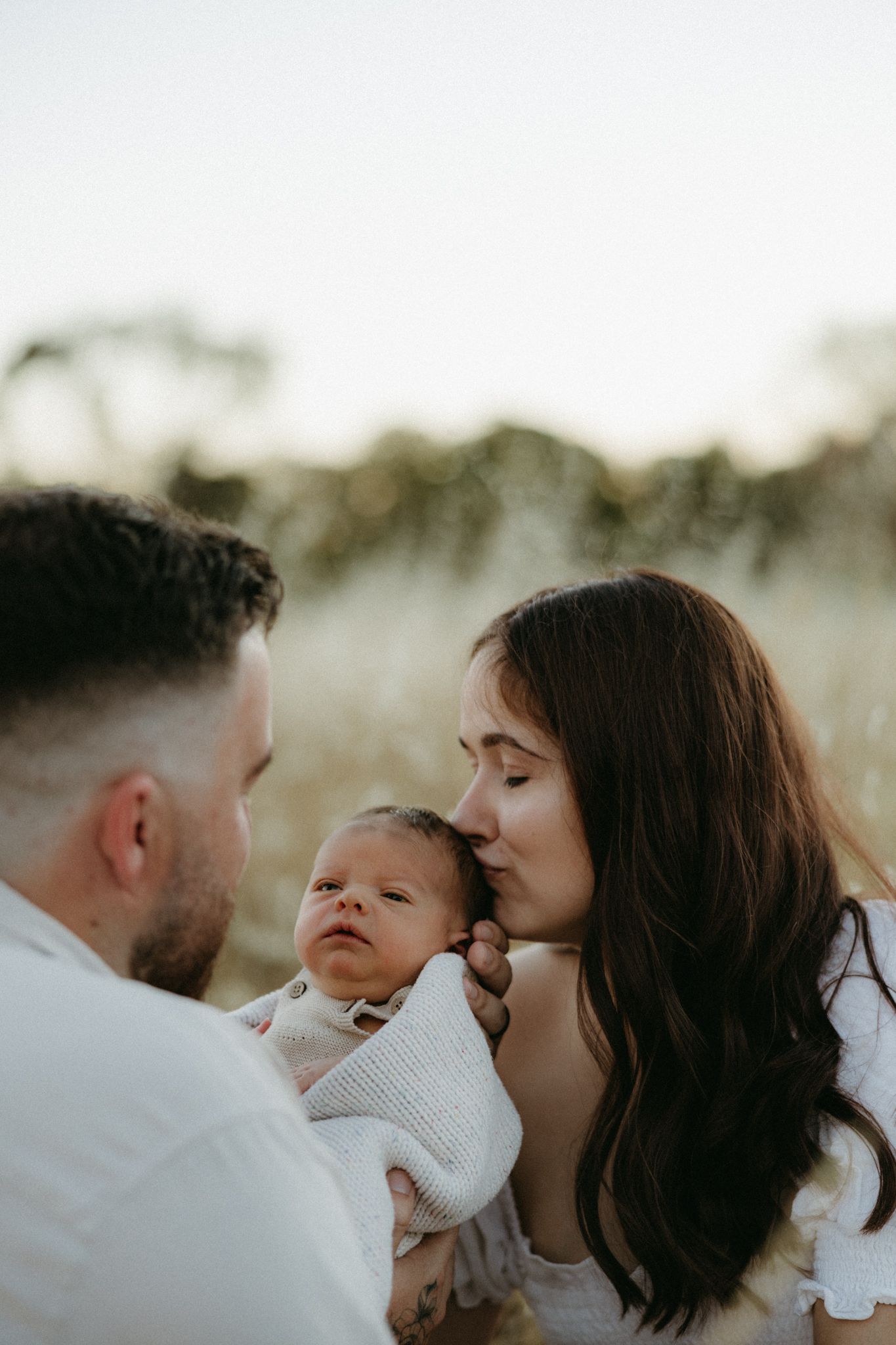 A couple shares a tender moment kissing their newborn baby outdoors in a golden field.
