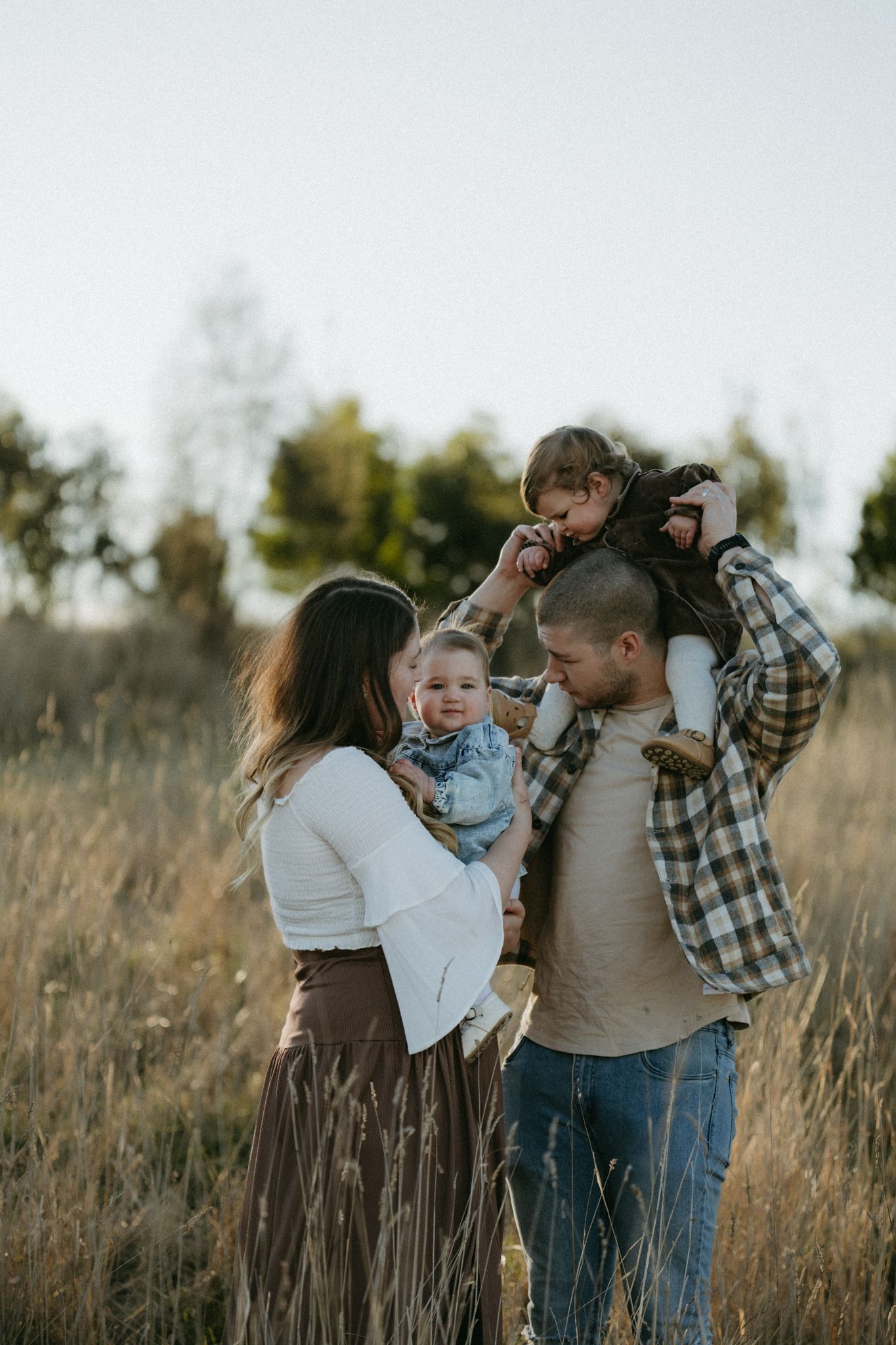 Family of four enjoying a golden hour outdoor photo session in a field, parents holding two young children.