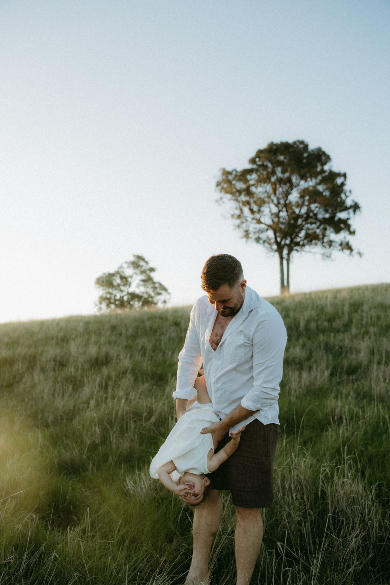 A father holds his baby upside down playfully in a sunlit green field with a lone tree on the hill behind him.