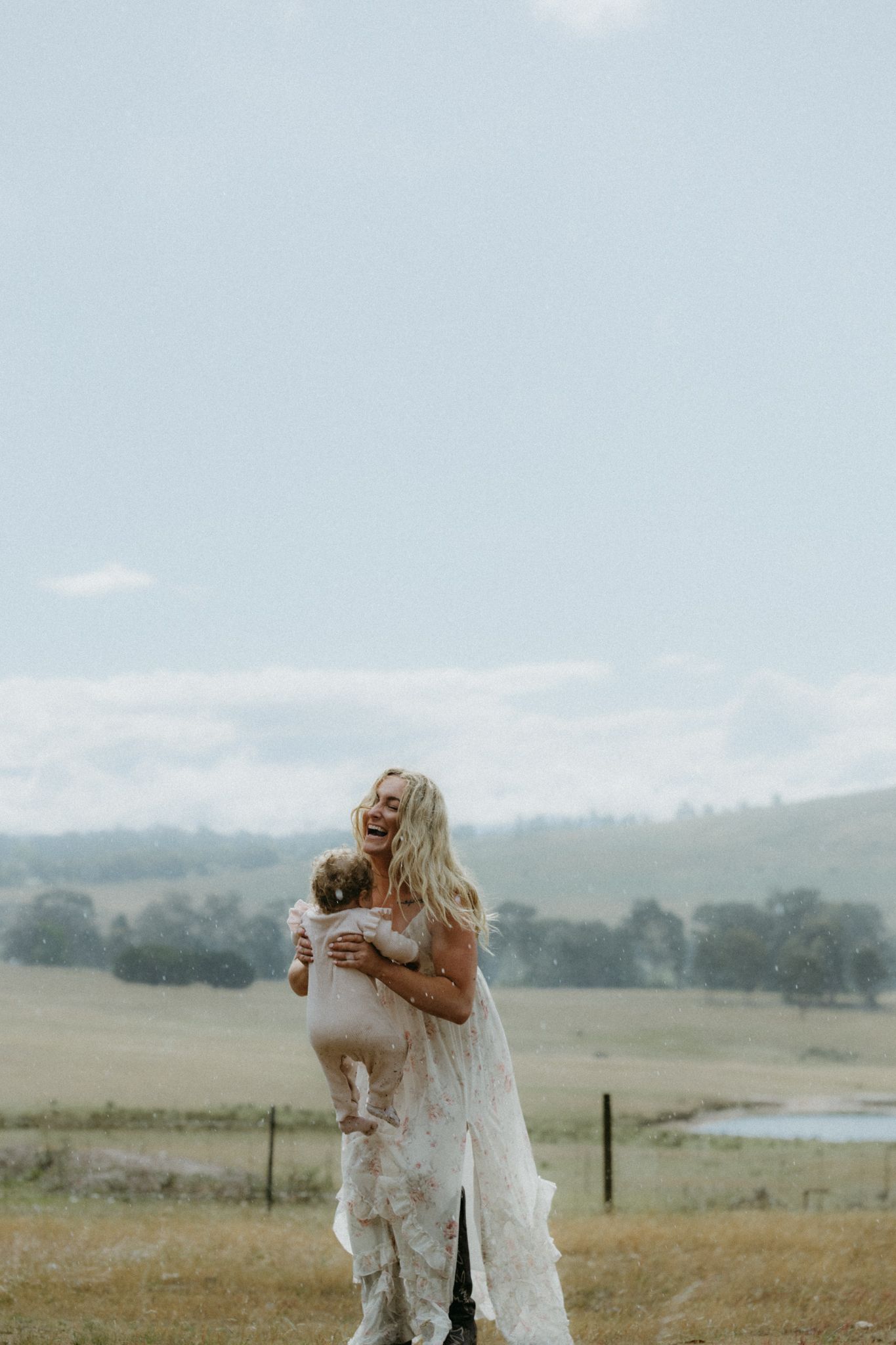 Bride in lace wedding dress laughing while holding a baby in a scenic rural field with green hills.