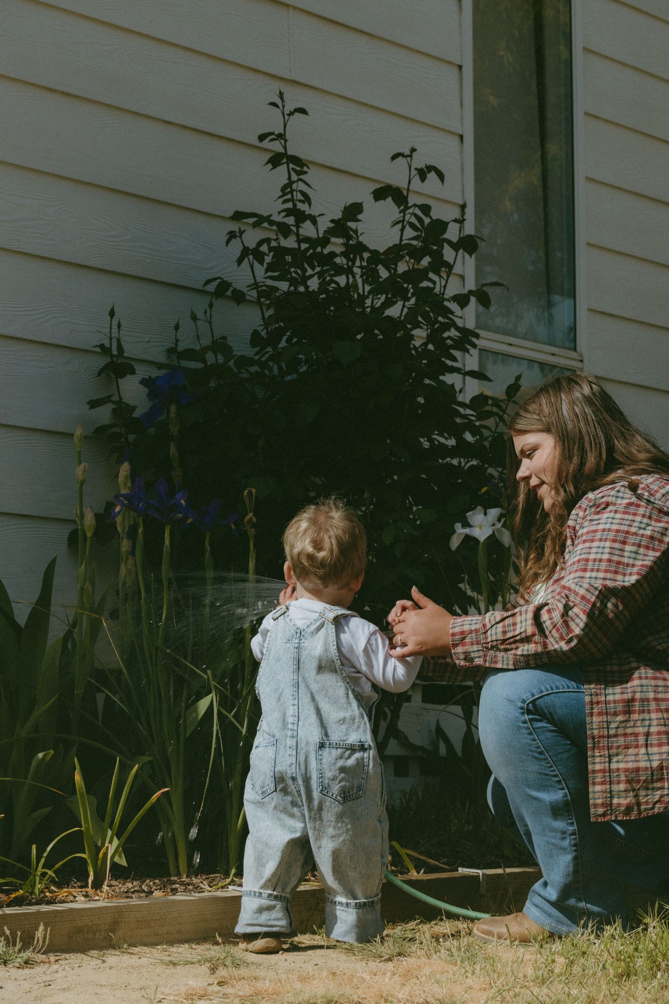 A mother crouches down smiling at her toddler in denim overalls taking first steps in a garden beside a house.