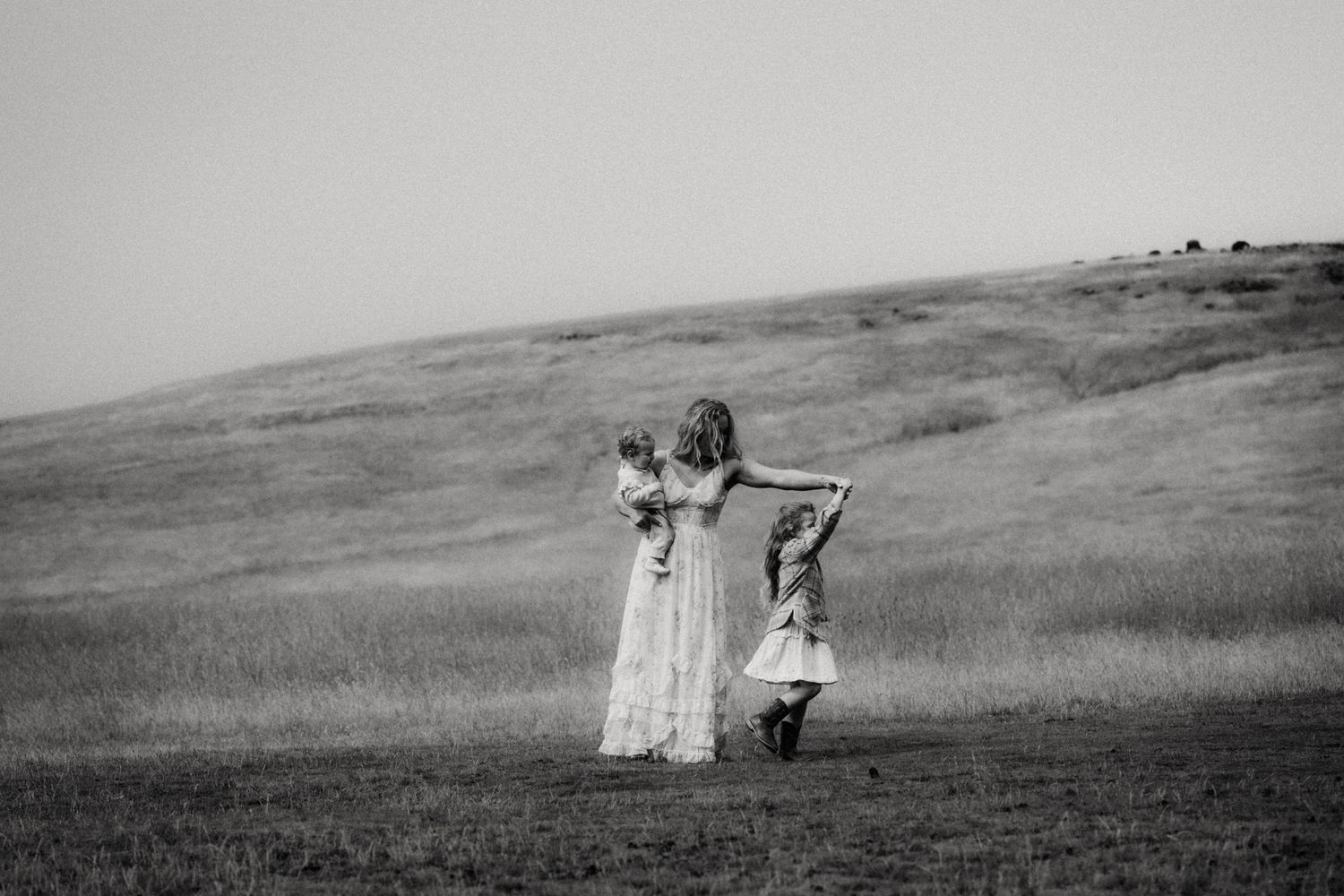 Black and white photo of a mother in a flowing dress walking with two young children across a grassy hillside meadow.