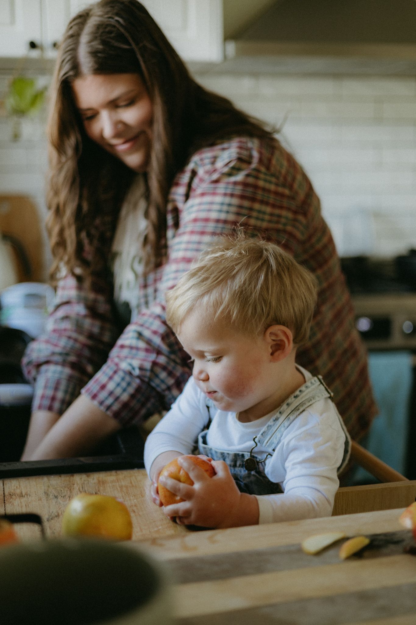 A smiling woman in plaid shirt watches a toddler in denim overalls holding an orange on a wooden tray in a kitchen.