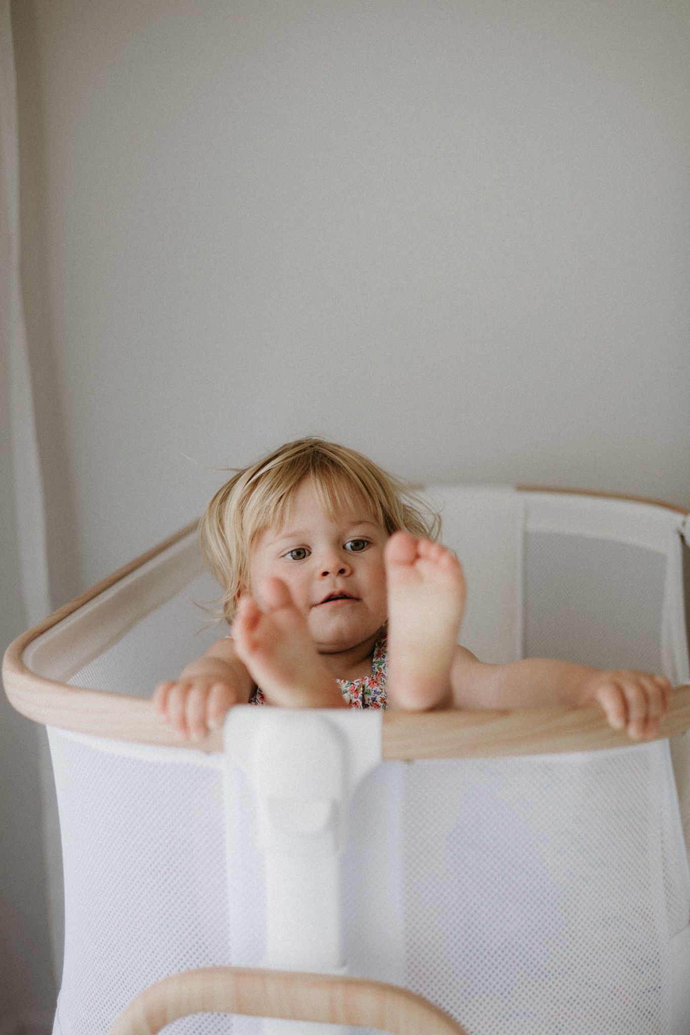 Toddler girl with blonde hair standing in a white wooden crib, showing her bare feet over the rail.
