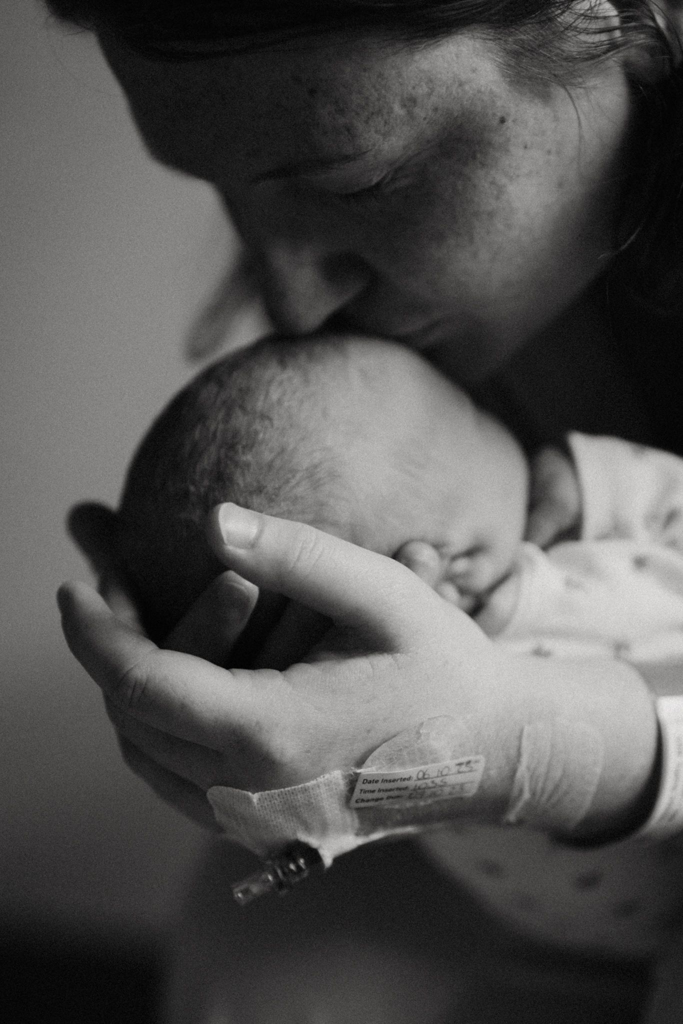 Black and white photo of a new mother tenderly kissing her newborn baby while holding them in a hospital room.