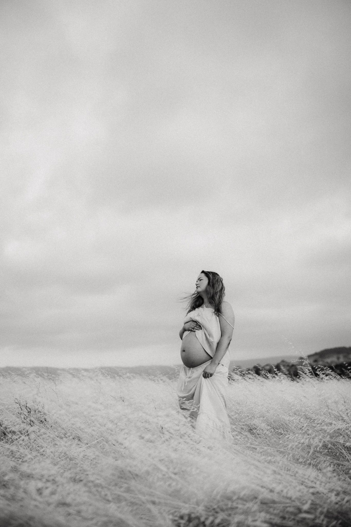 Black and white maternity photo of pregnant woman in flowing dress standing in field of tall grass under cloudy sky.