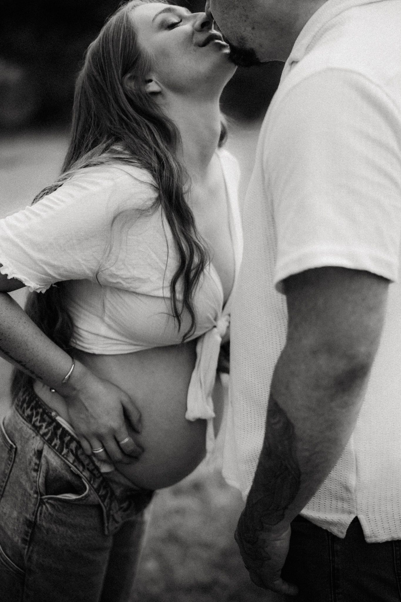 Black and white maternity photo of pregnant couple sharing a kiss, woman cradling baby bump in jeans and white top.